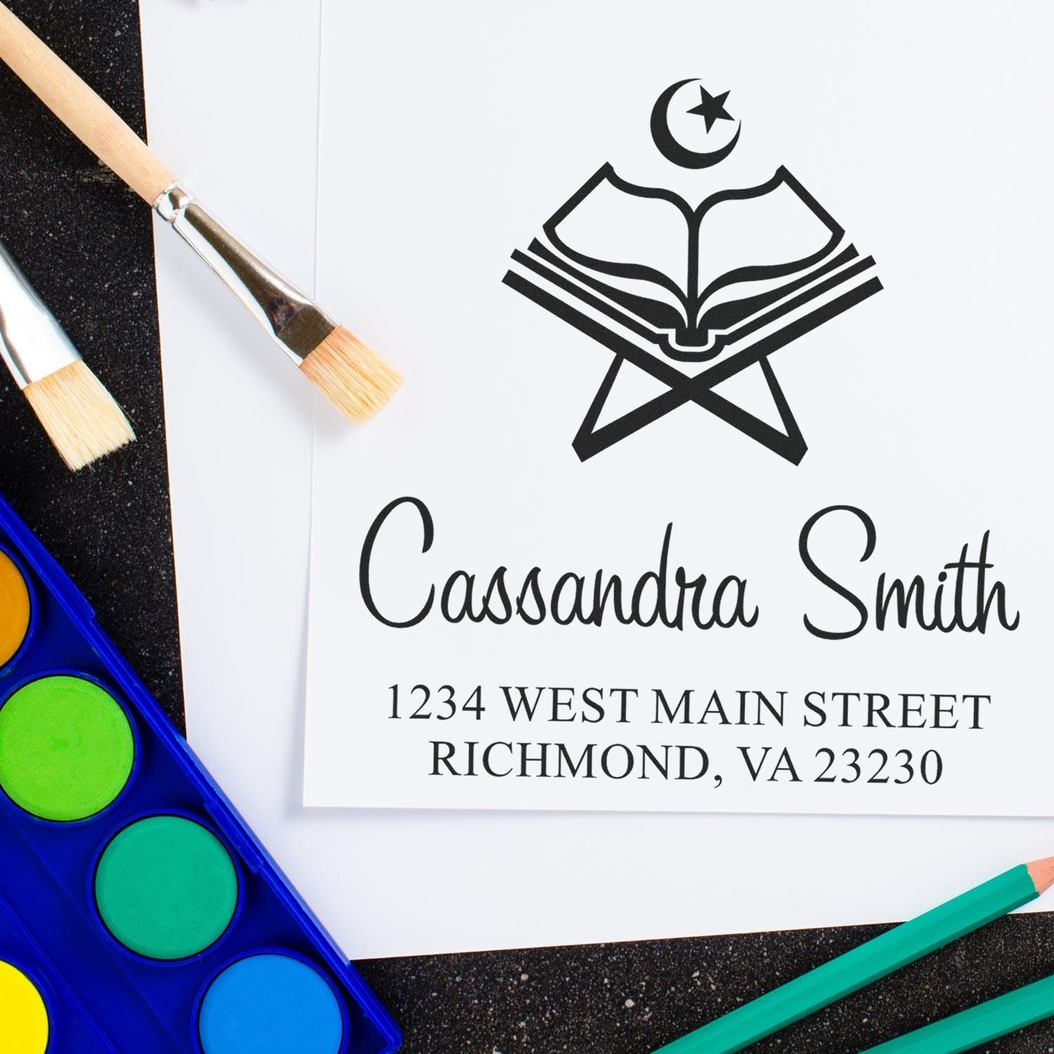 Custom stamp and seal with black book-and-crescent logo, Cassandra Smith name, and address on white paper beside paintbrushes and watercolors.