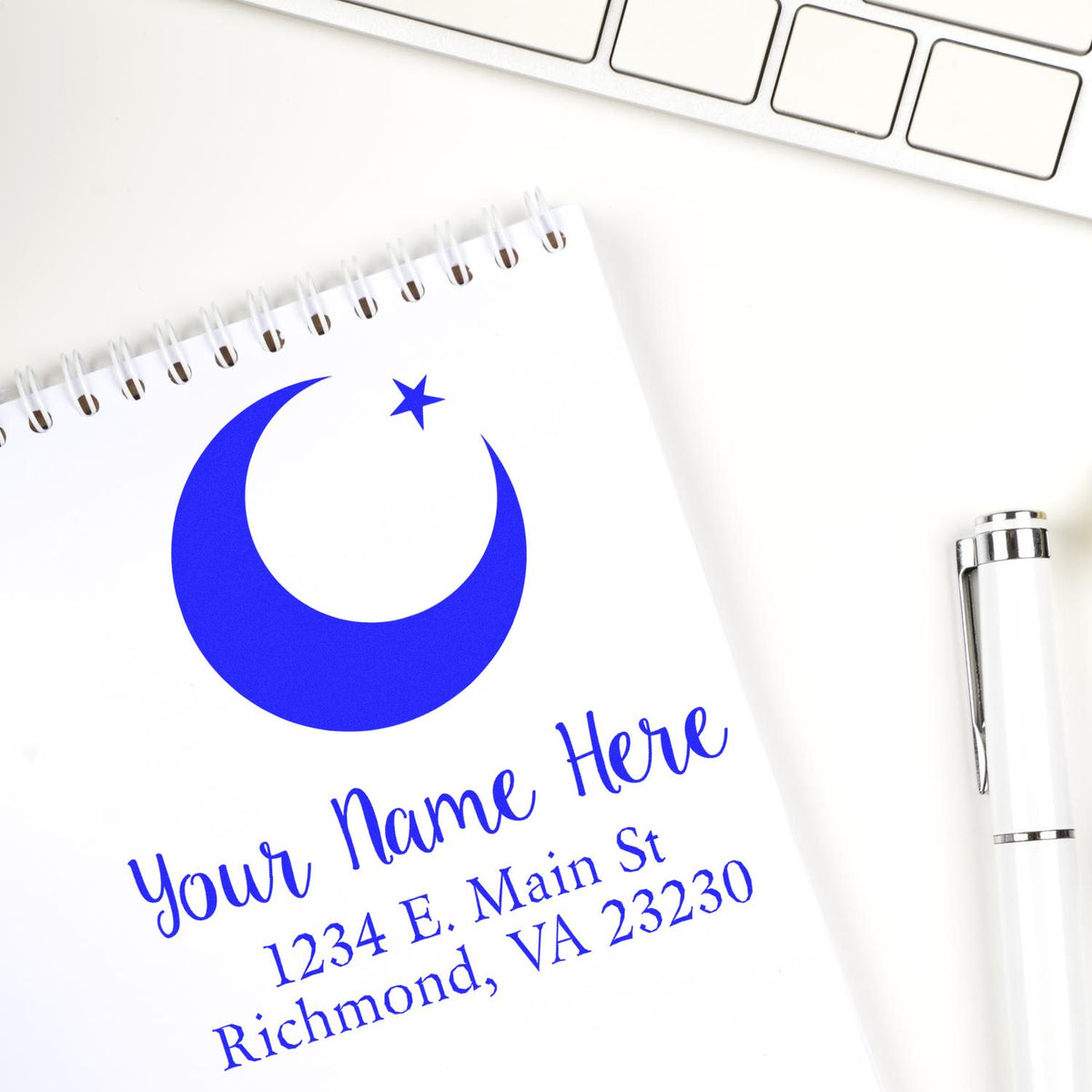 Blue custom stamp seal with crescent moon and star logo, name and address on a white notepad beside a silver pen.