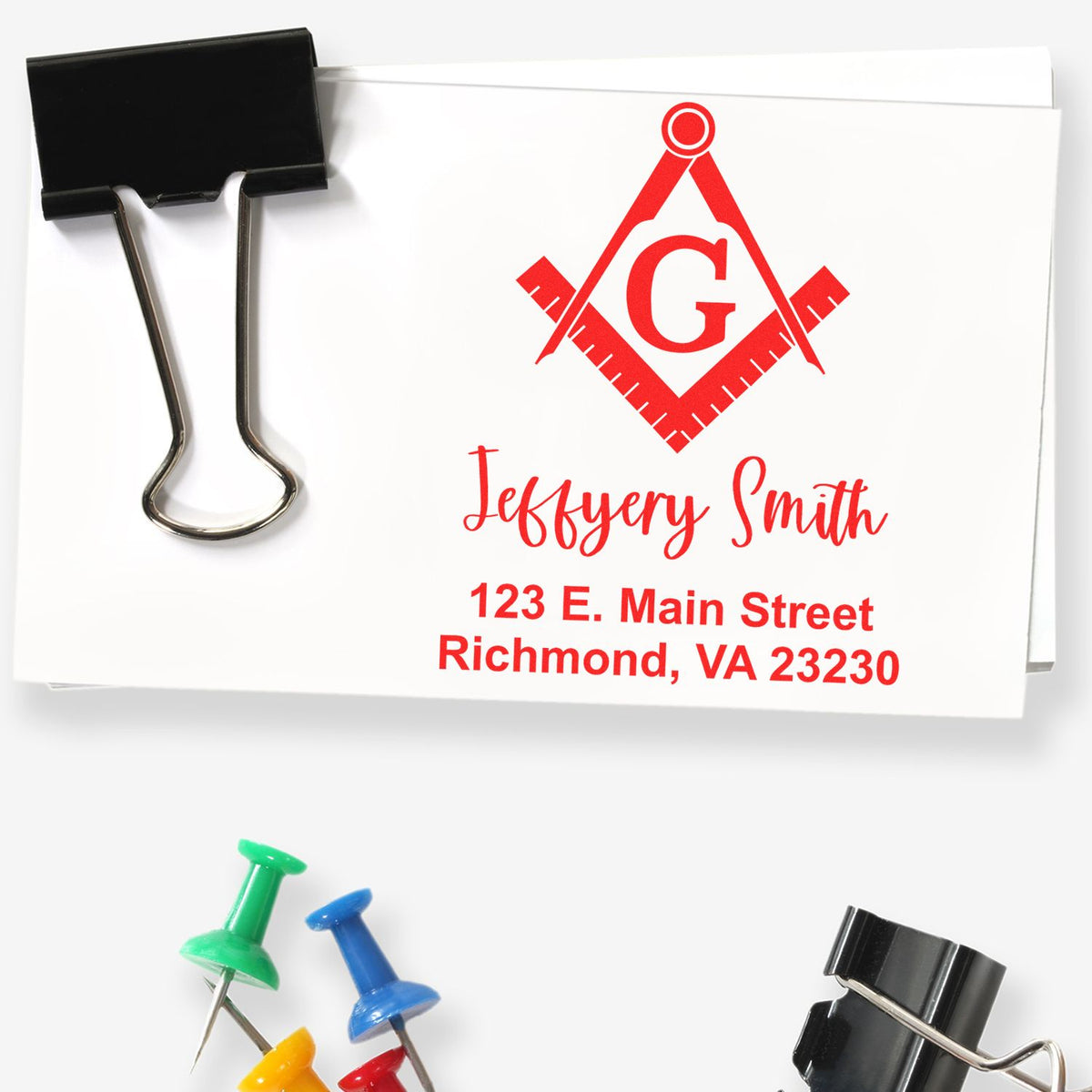 White custom stamp and seal cards with red logo, name and address, clipped by a black binder clip and colorful push pins.