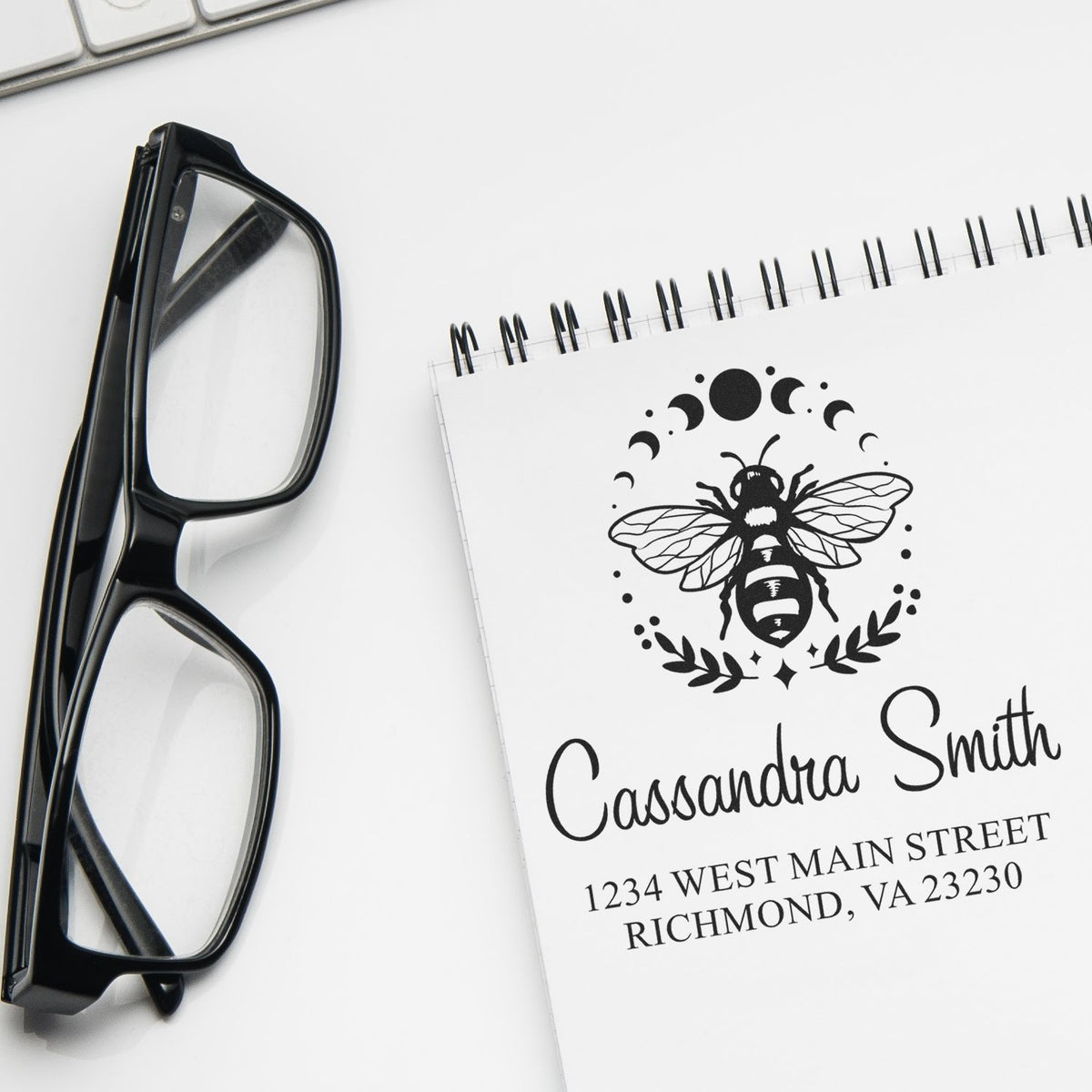 Black eyeglasses beside a white spiral notebook with a custom bee seal stamp, name, and address in black.