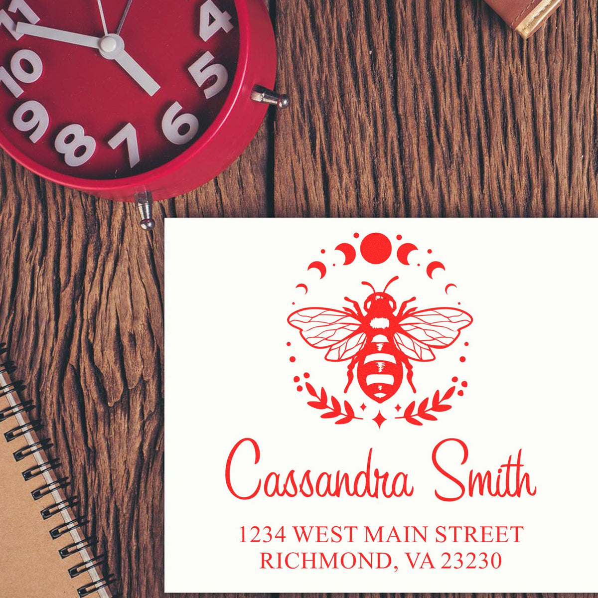 Red custom bee address stamp and seal on white card, with woodgrain desk and red clock