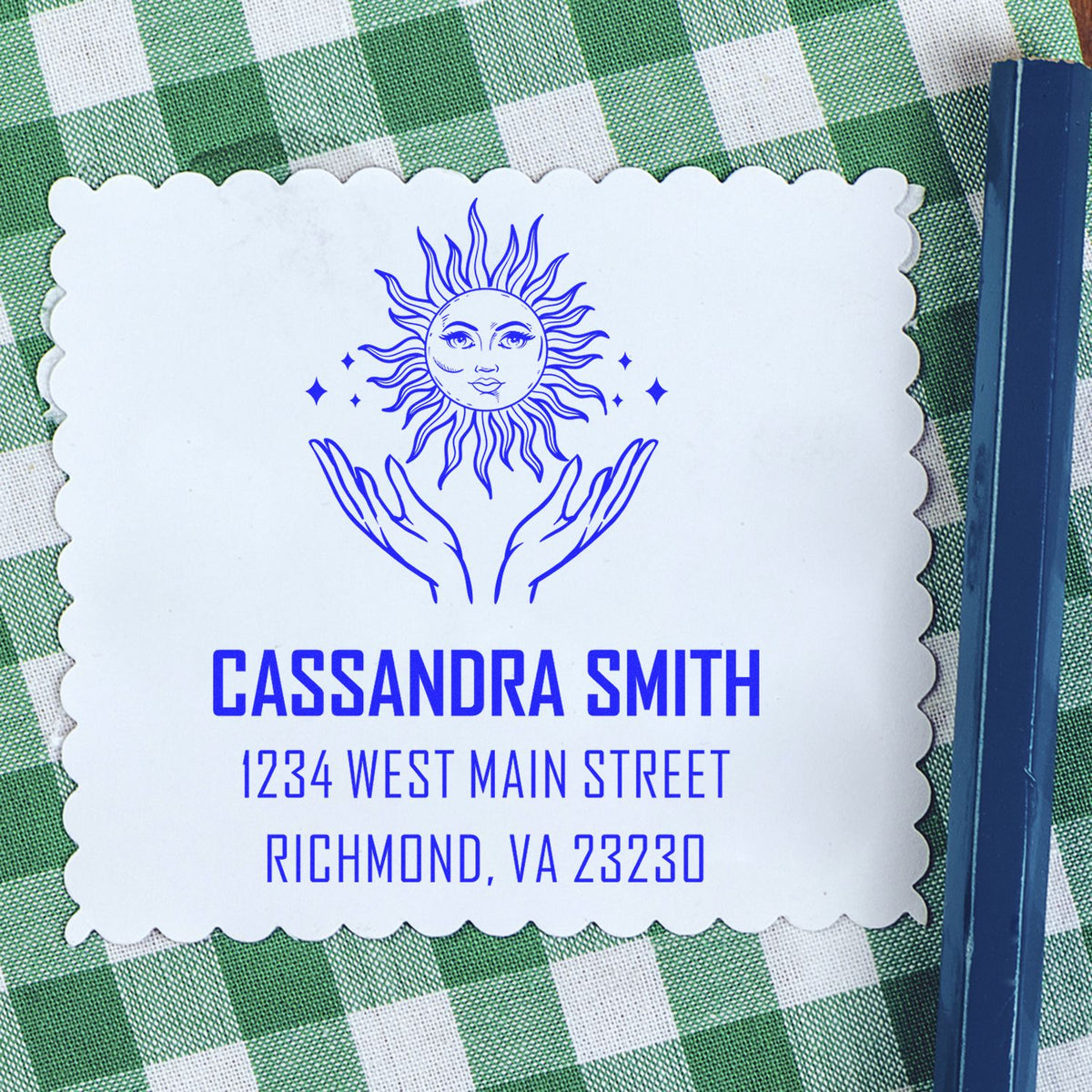Blue custom address stamp and seal with sun-and-hands design on white scalloped card over green gingham cloth.
