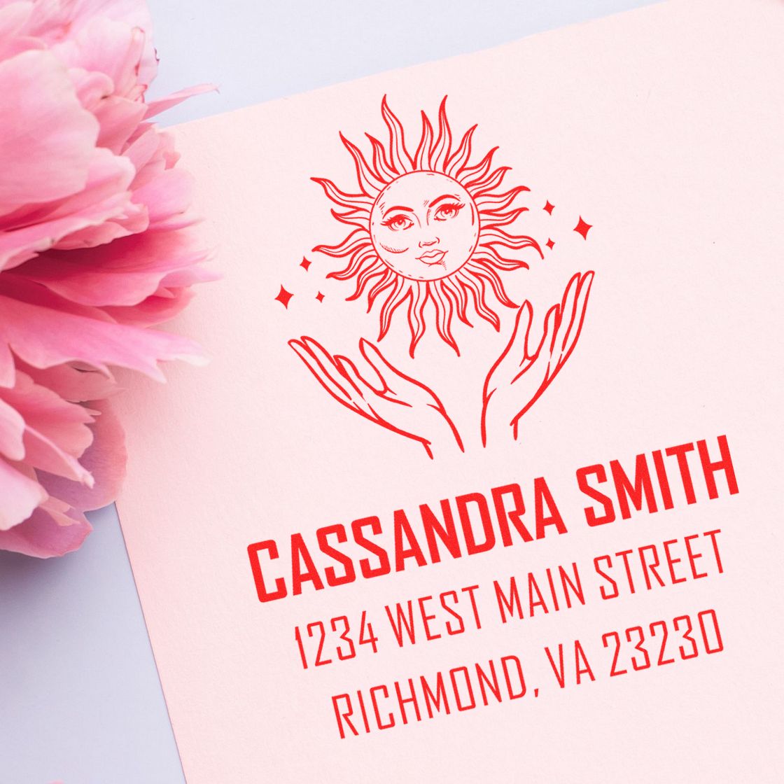 Red personalized stamp and seal with sun-and-hands design, name and address on pale pink paper beside pink peonies.