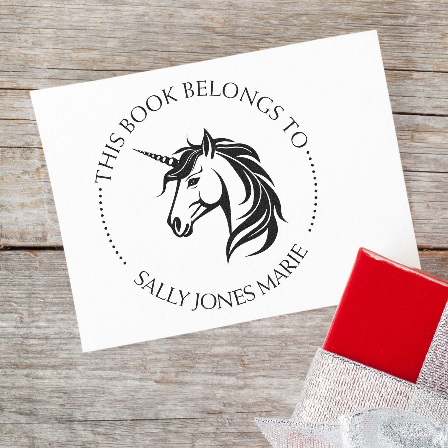 Custom black unicorn book stamp on white card reading This Book Belongs to Sally Jones Marie, with red stamp seal on wood background.