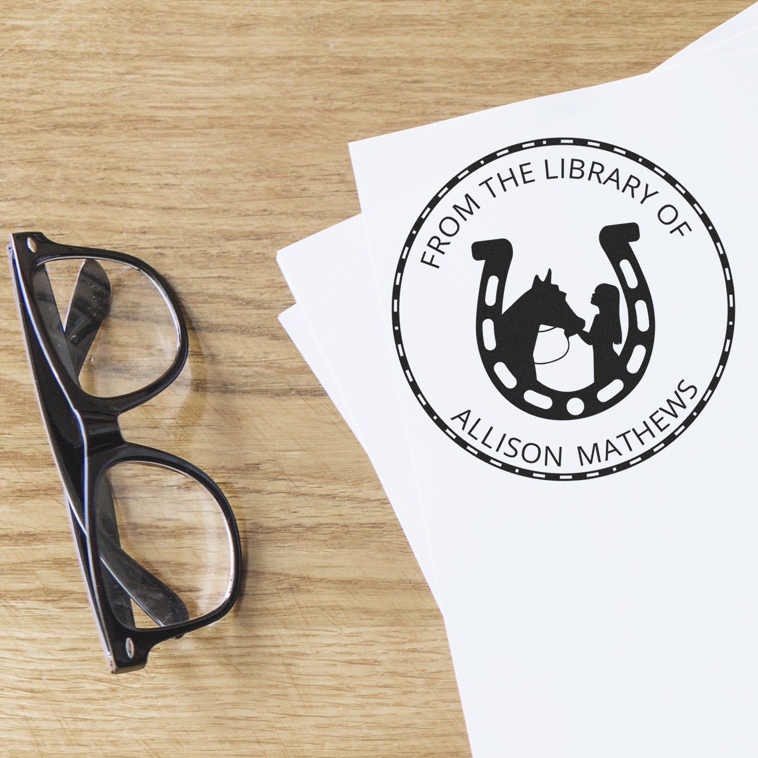 Black custom library stamp and seal with horseshoe and horse design on white paper, beside black eyeglasses on a wood desk