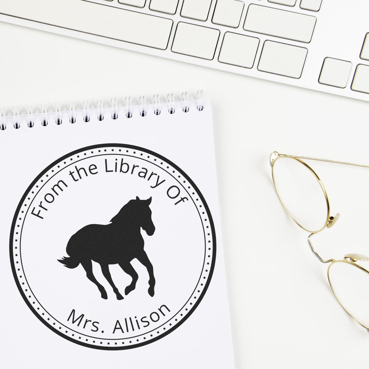 Black circular custom horse library stamp seal reading From the Library of Mrs. Allison on a white notebook, with keyboard and gold glasses.