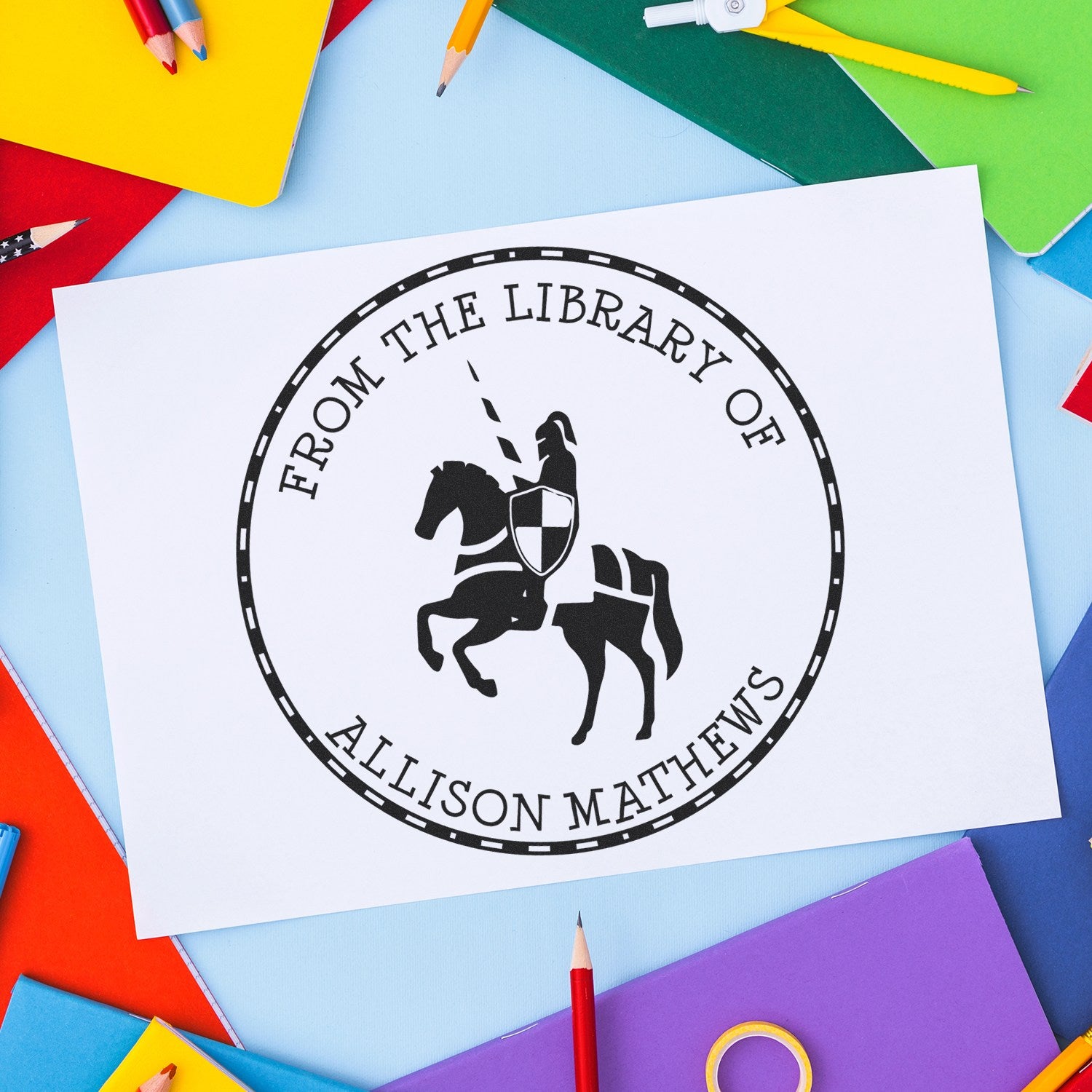 Black custom library seal stamp for Allison Mathews with knight on horse, on white paper amid colorful pencils and notebooks.