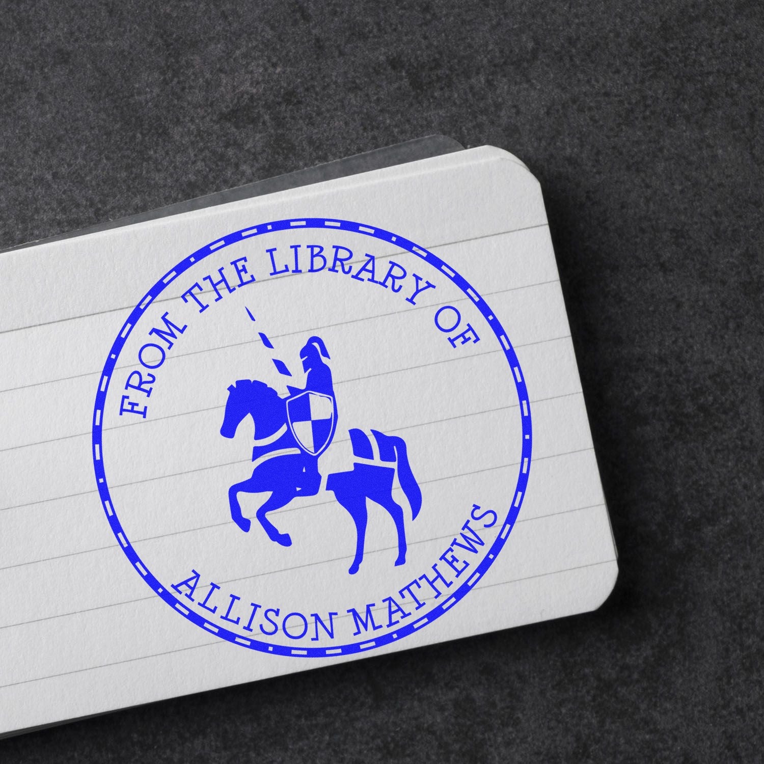Blue circular library seal stamp with mounted knight and shield on white lined card over a dark background.