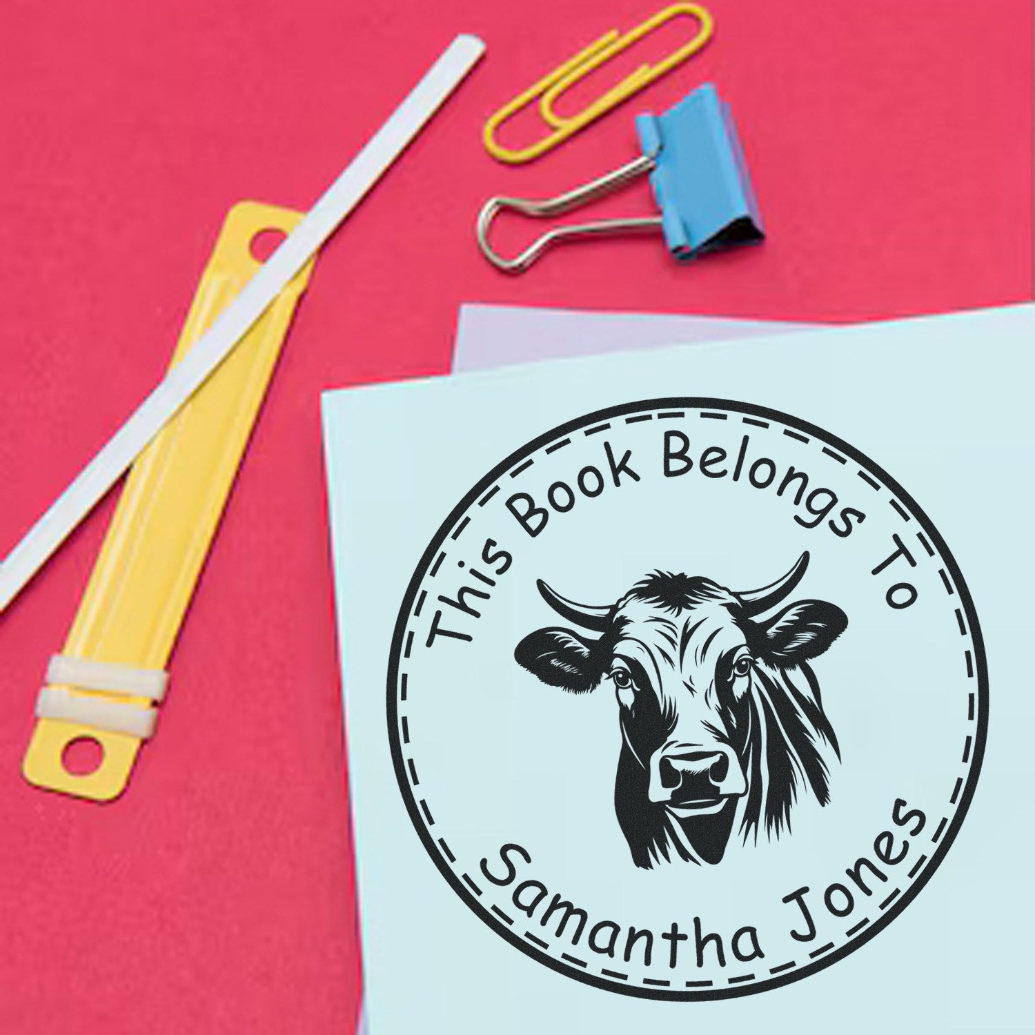 Light blue custom bookplate stamp seal with cow graphic and This Book Belongs To Samantha Jones on pink background