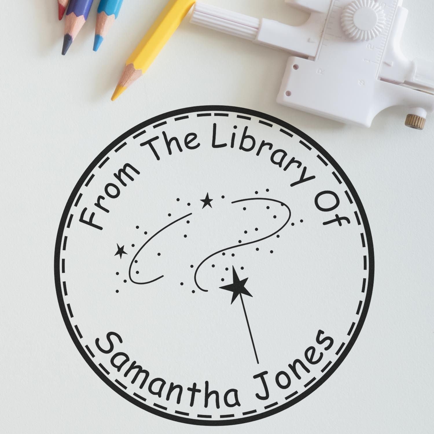 Black circular library stamp seal reading From The Library Of Samantha Jones on white paper, with colored pencils and a white embossing tool.