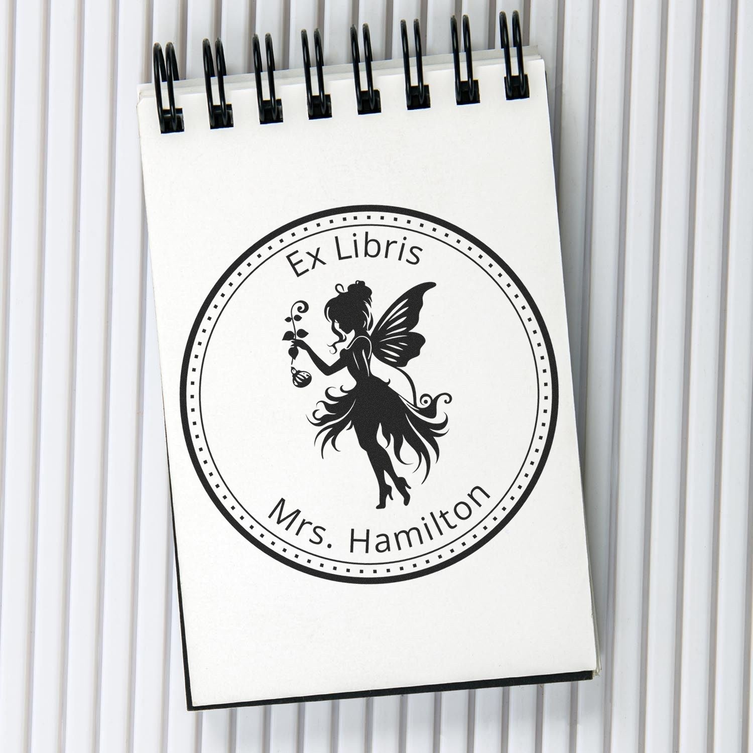Black ex libris book stamp on white spiral notepad, featuring a fairy silhouette and Mrs. Hamilton in a circular seal.