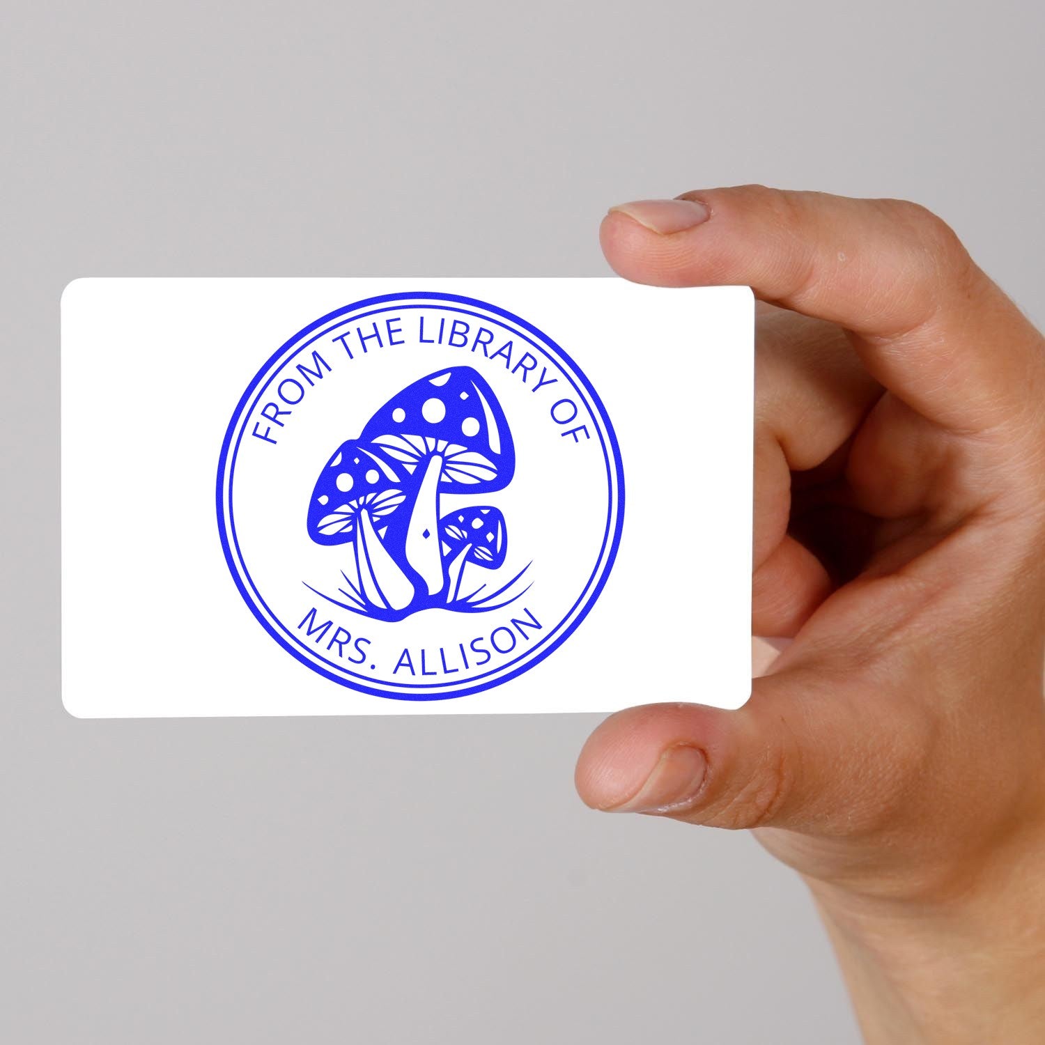 Blue custom round library stamp seal with mushroom design and Mrs. Allison text, held on a white card by hand.