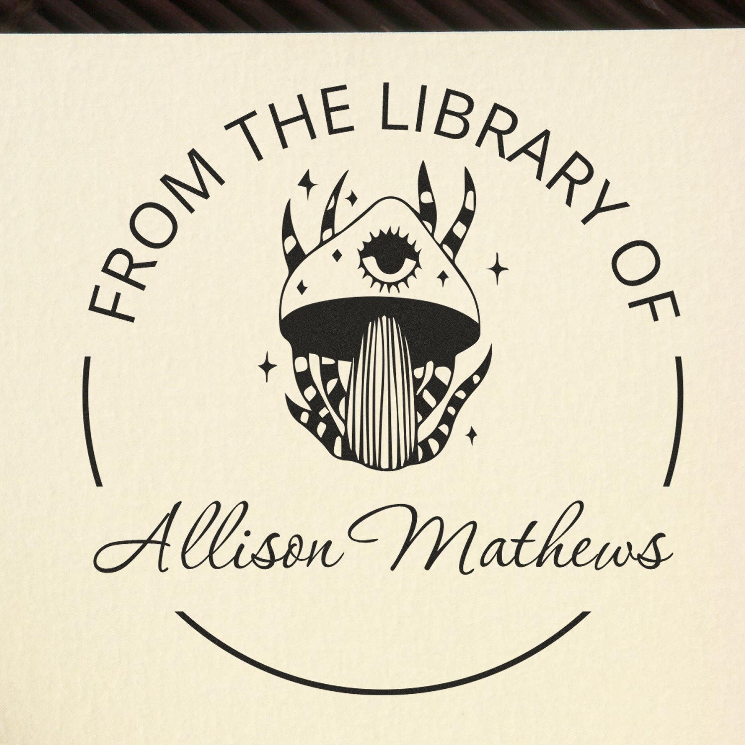 Black circular custom library stamp seal with horned mushroom emblem, stars, and Allison Mathews on cream background.