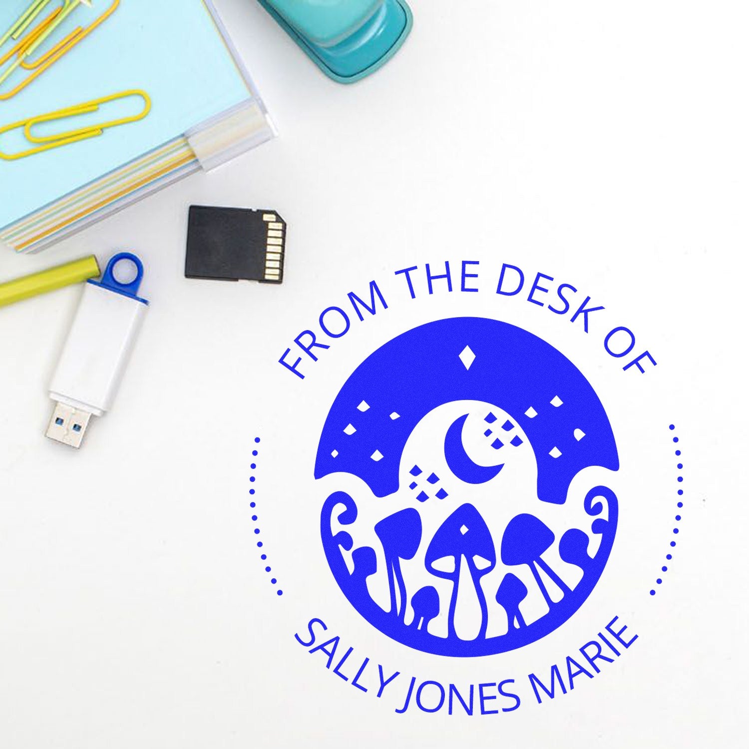 Blue custom desk seal stamp with mushrooms and crescent moon, beside USB drive, paper clips, and memory card on white background.