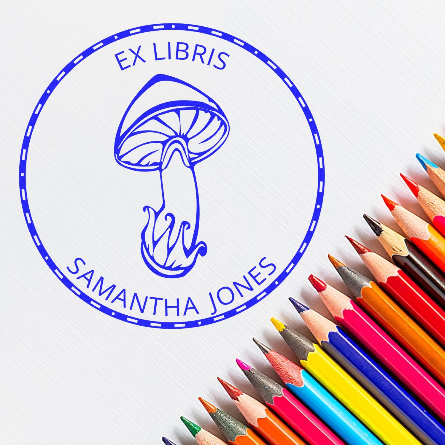 Blue EX LIBRIS mushroom book stamp seal with Samantha Jones text, beside colorful pencils on a white background.