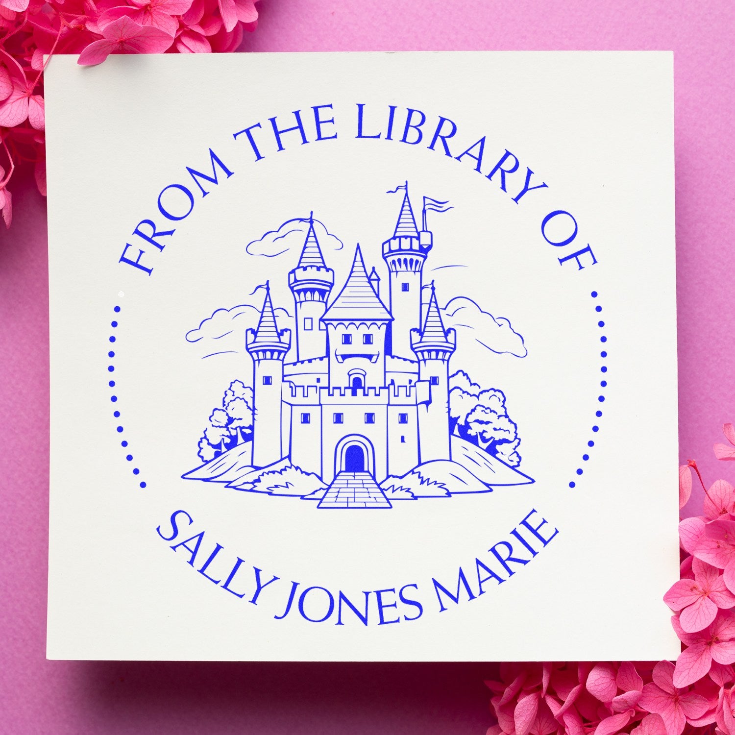 Blue custom library stamp seal with castle, reading FROM THE LIBRARY OF SALLY JONES MARIE, on white paper with pink flowers.