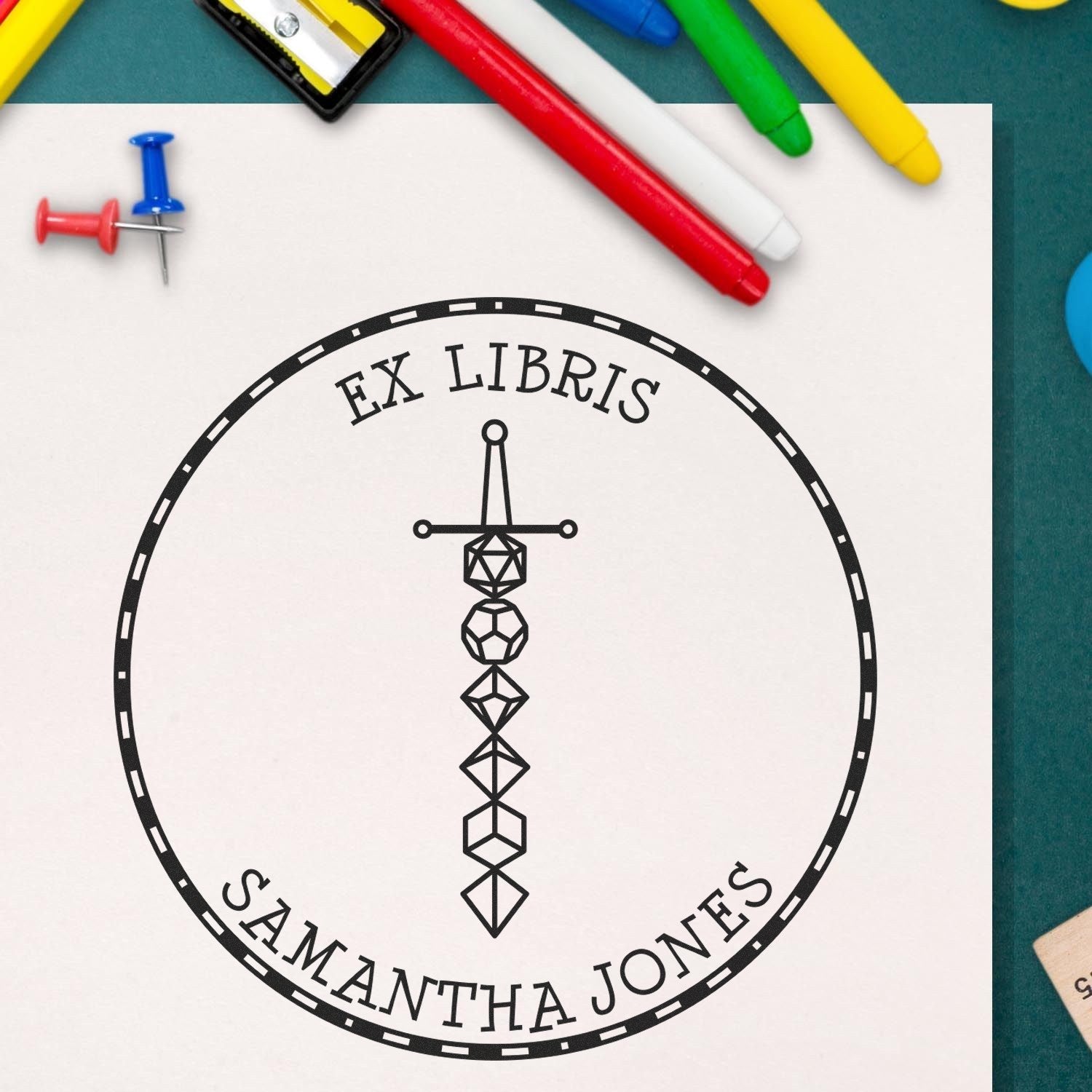 Black custom ex libris bookplate stamp seal with geometric sword emblem and Samantha Jones text on white paper, with colorful markers.