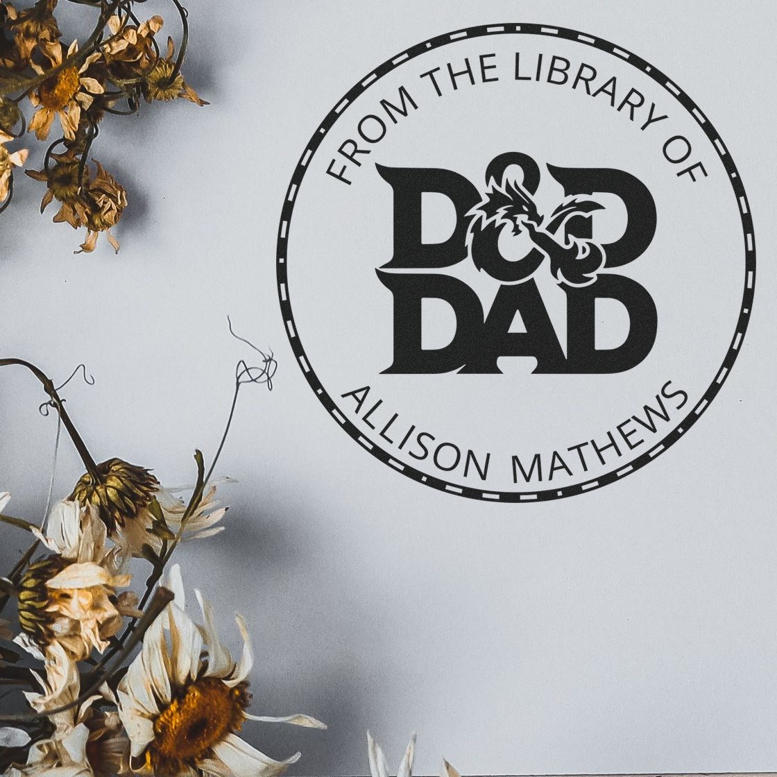 Black custom library stamp seal reading "From the Library of Dad" with Allison Mathews, on white background and dried flowers.
