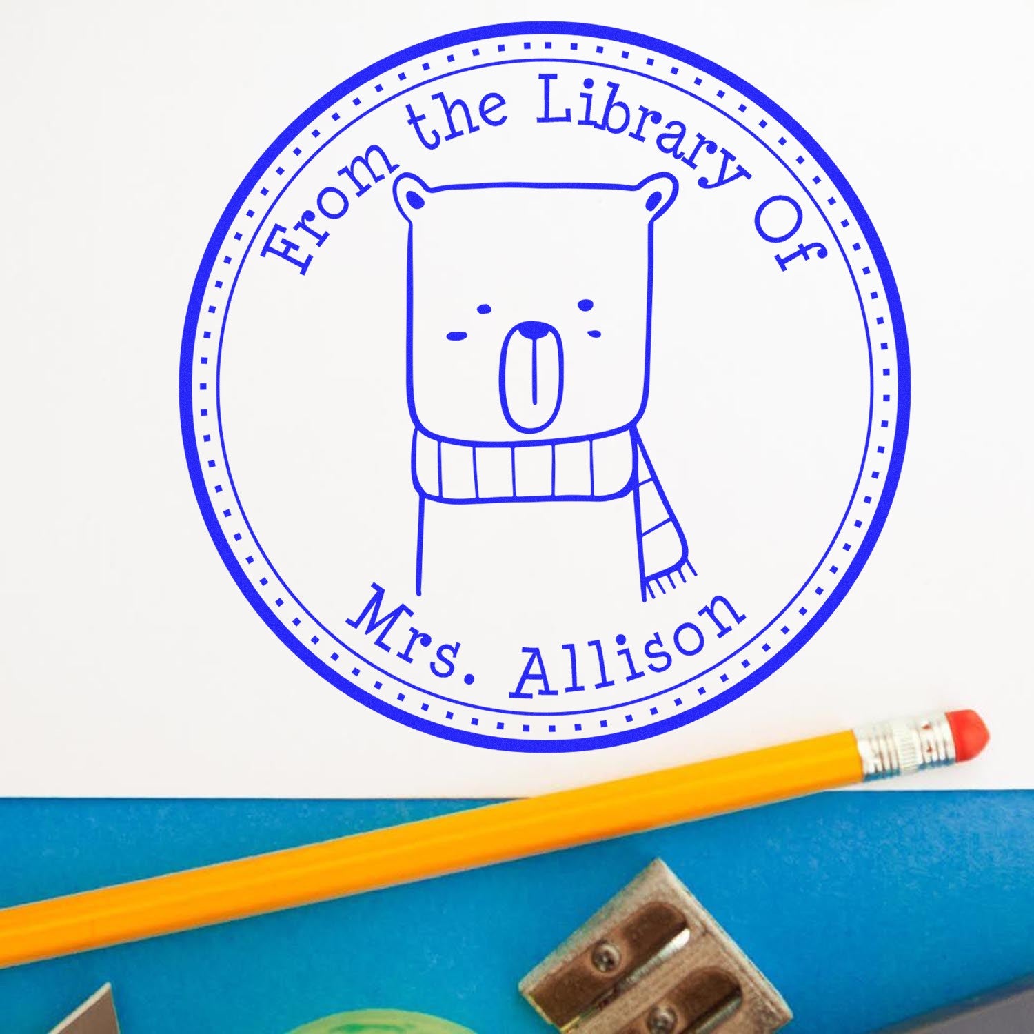 Image of a custom mailing stamp with a wood handle, featuring a BounceTail cartoon animal design. The personalized book design stamp reads "From the Library Of Mrs. Allison" in blue ink, next to a pencil and eraser.