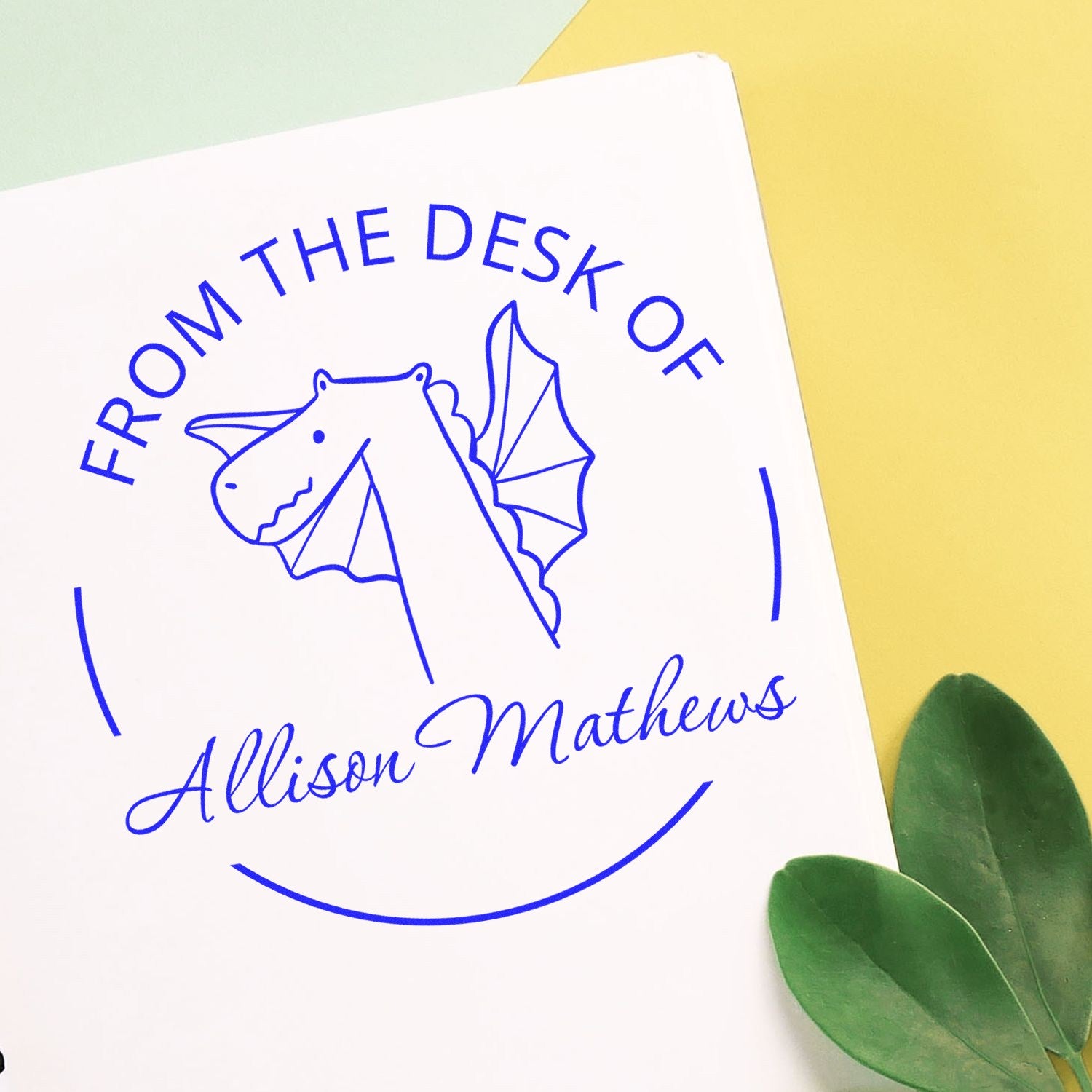 Slim Pre-Inked SnuggleSnout Cartoon Animal Custom Book Identification Stamp on paper, featuring a dragon illustration with text "From the Desk of Allison Mathews." Customized Mailing Stamper design on a yellow and white background with green leaves.