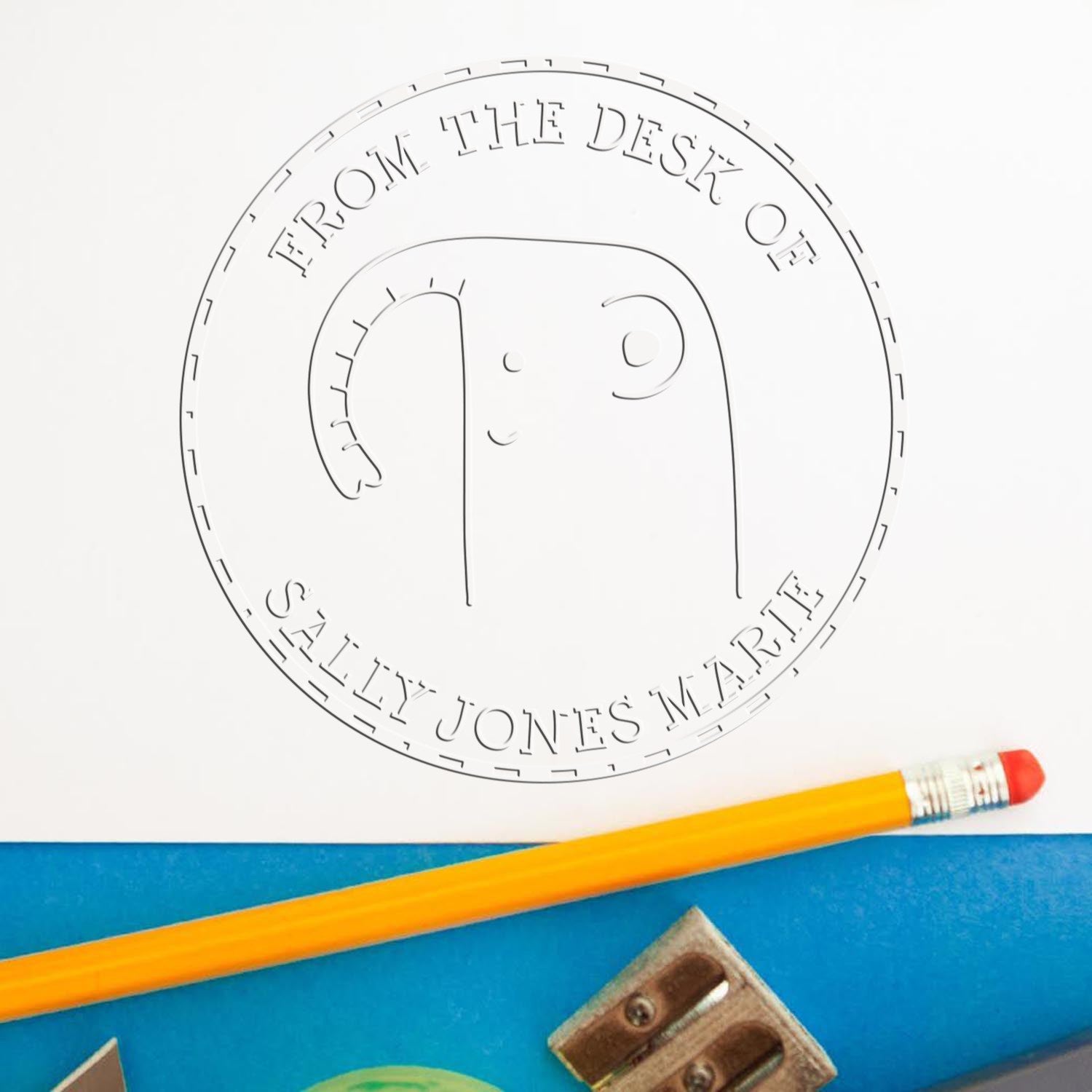 A GiggleWiggle cartoon animal hybrid design is embossed on paper using a customized library return embosser. The custom-made mailing stamp reads "From the Desk of Sally Jones Marie" with a pencil and sharpener nearby.