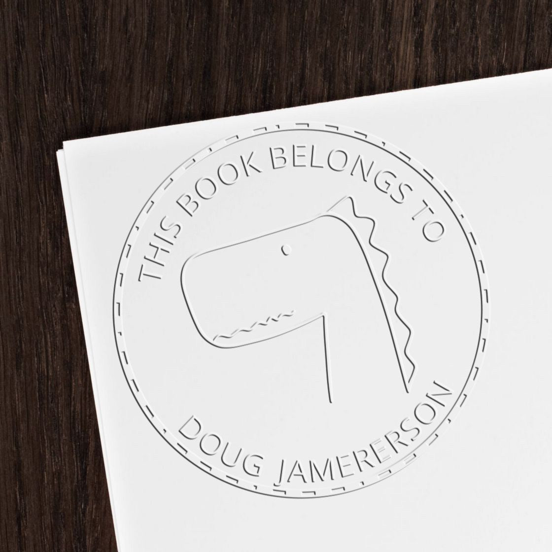 A Soft JellyBeanJump cartoon animal design is embossed on a white paper using a custom-made book marking embossing seal. The seal reads This Book Belongs To Doug Jamerson. The image showcases a custom mailing stamper on a dark wooden surface.