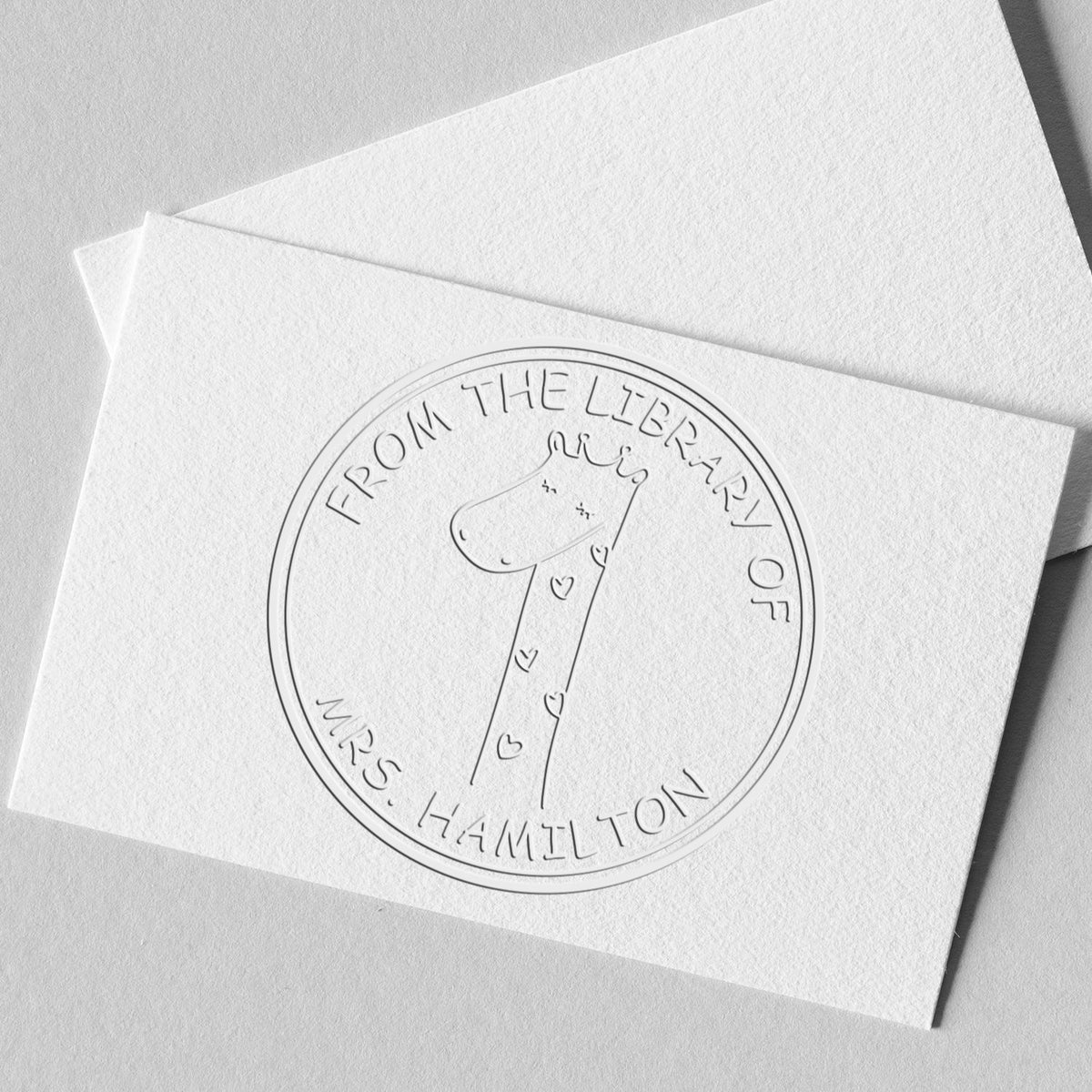 Embossed card with a SquiggleSquash cartoon animal hybrid design, featuring a giraffe with hearts. Text reads "From the Library of Mrs. Hamilton." Includes a customizable new address stamper and library return embossing seal.