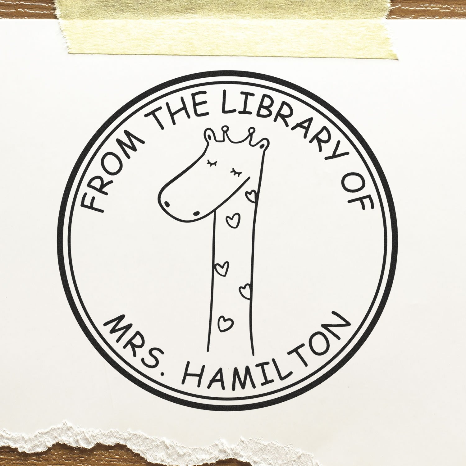 A SquiggleSquash cartoon animal stamp with a giraffe design, labeled "From The Private Library Of Mrs. Hamilton," is shown. This pre-inked, customizable stamp is ideal for a custom address stamper. The stamp is on white paper with torn edges and taped at the top.