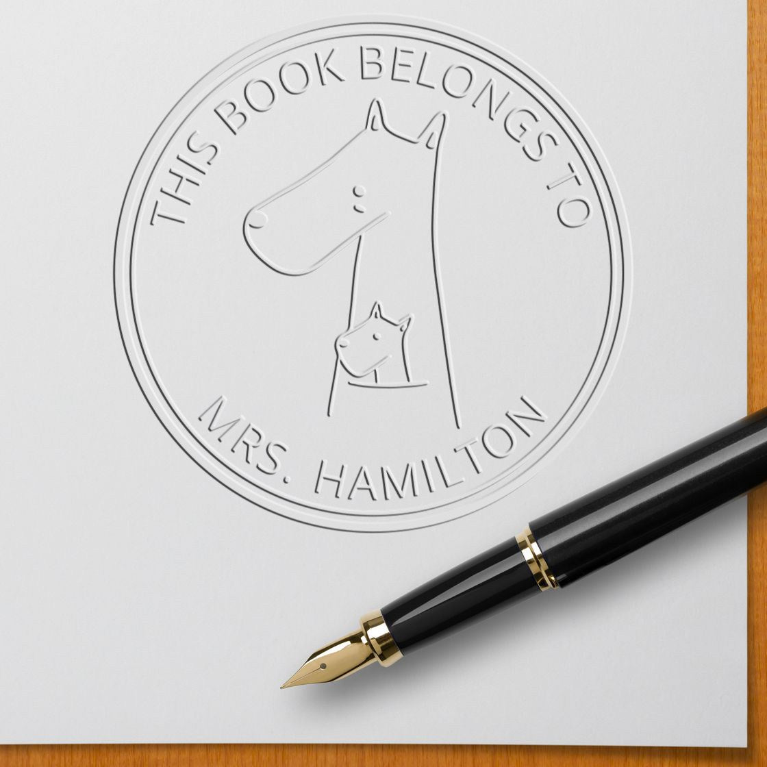 Embossed seal on paper featuring a MuddleMoo cartoon animal hybrid, customized from the bookshelf of embosser. Includes text "This Book Belongs To Mrs. Hamilton." A fountain pen lies nearby. Personalizable new address stamper concept.