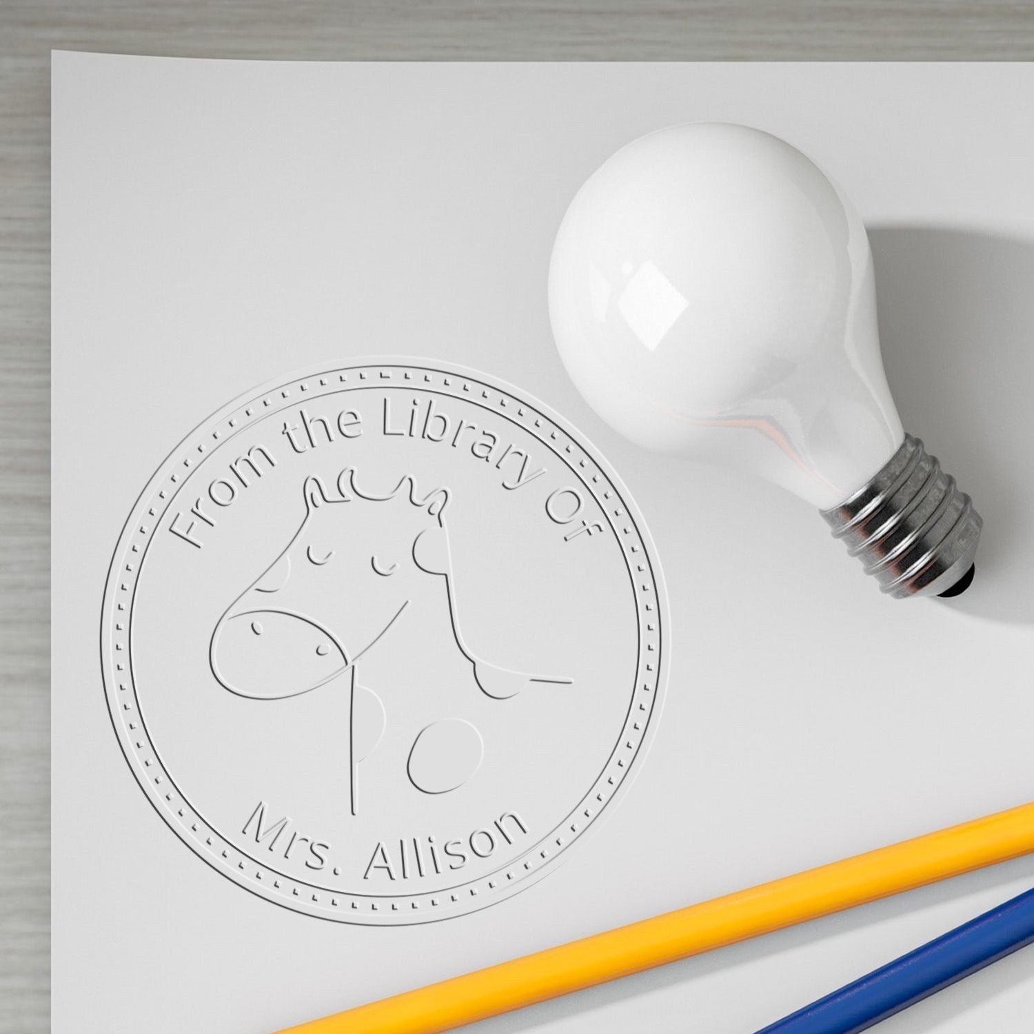 A BoopSnoot cartoon animal design is embossed on a handheld personalized ex libris book seal. The custom address stamper features a cute animal face with the text "From the Library Of Mrs. Allison" on a white paper, next to a light bulb and pencils.