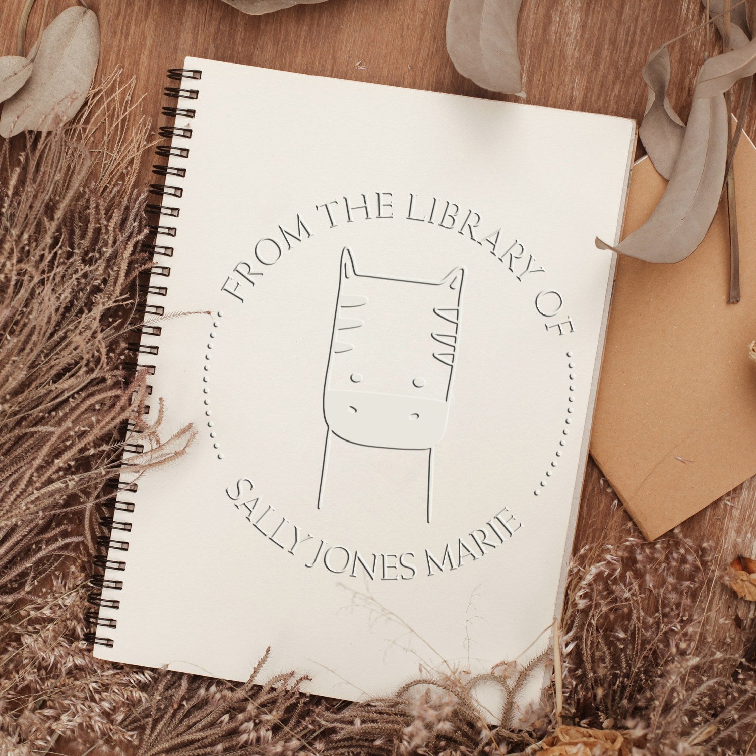 Cartoon animal design on a personalized ex libris book embossing seal, labeled "From the Library of Sally Jones Marie." The PuzzlePounce seal is on a spiral notebook, surrounded by dried leaves. Custom address stamp not visible.