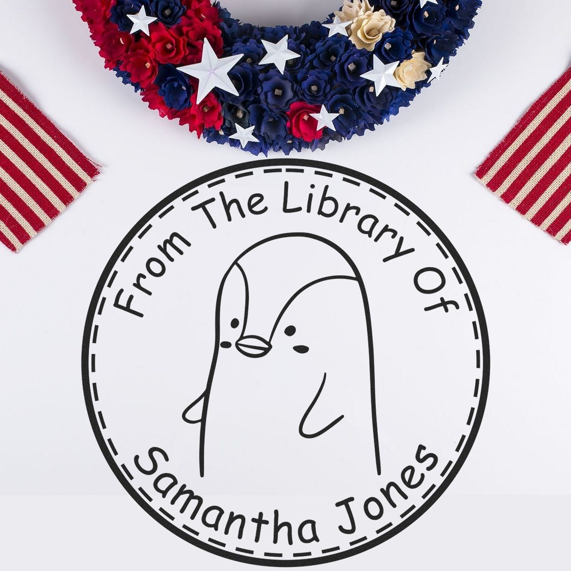 Slim Pre-Inked PebbleWobble Cartoon Animal Custom From The Bookshelf Of Stamp displayed on a white surface. The stamp features a cute penguin design with text "From The Library Of Samantha Jones." Nearby are patriotic decorations. Custom Home Stamper in use.