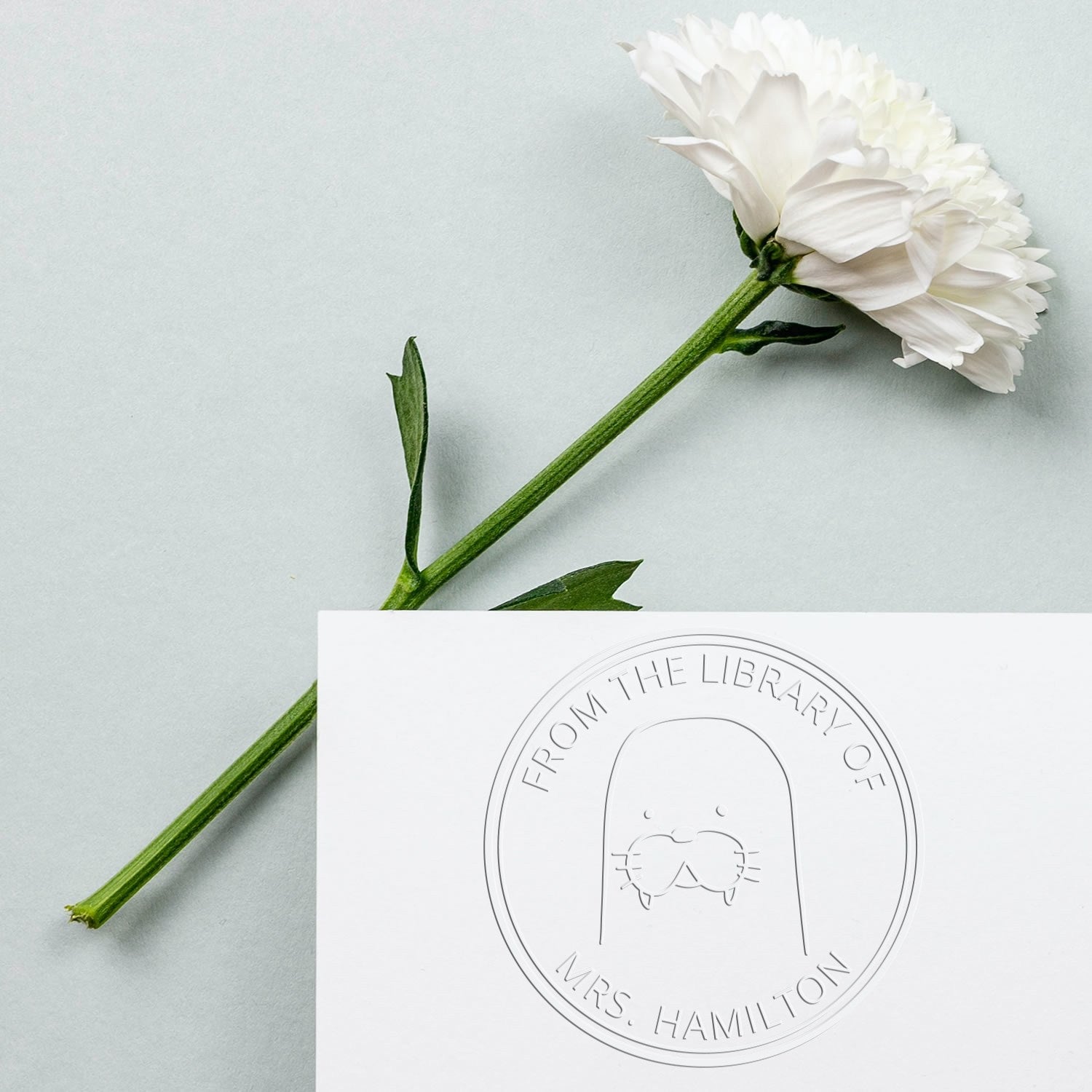A white card with a "From The Library Of" seal featuring a BumbleBristle cartoon animal. The seal is a customized new address stamper. A white flower lies beside the card on a light background.