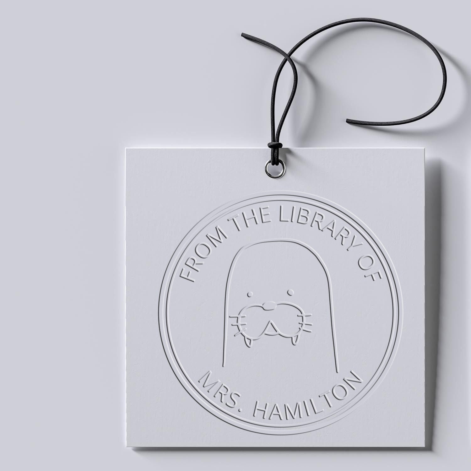 A white card features a BumbleBristle cartoon animal seal with the text "From the Library Of Mrs. Hamilton." The card is attached with a black string. This personalized item is a customized new address stamp.