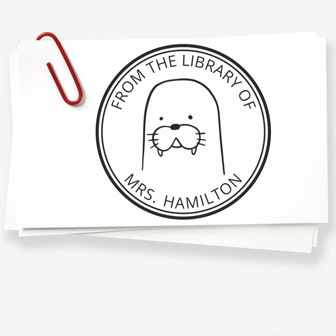 BumbleBristle Cartoon Animal Pre-Inked Customizable Ex Libris Design Stamp featuring a walrus illustration with "From the Library of Mrs. Hamilton" text. A Custom New Address Stamper is shown on a stack of cards with a red paperclip.
