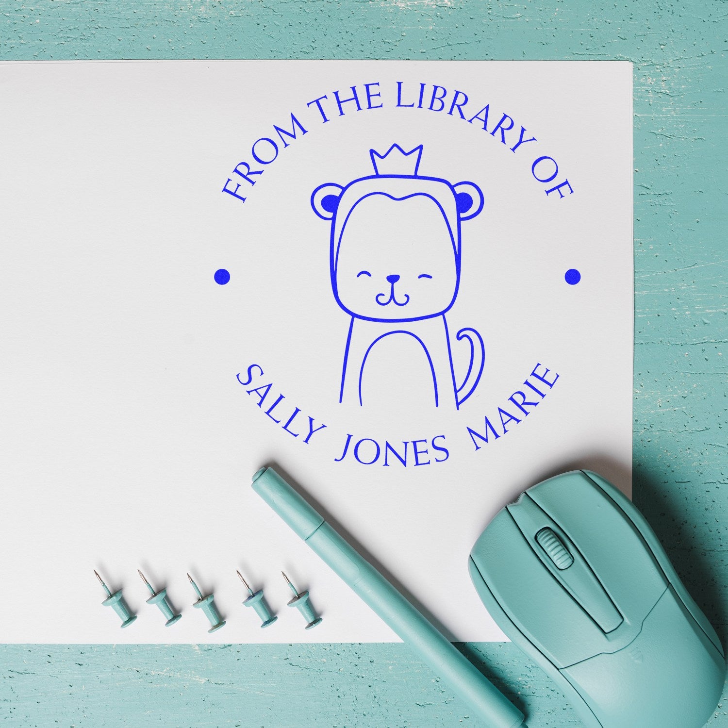 Slim Pre-Inked ScribbleScrabble Cartoon Animal Custom Library Stamp on white paper with a cute animal design and text "From the Library of Sally Jones Marie." Nearby are a teal pen, mouse, and push pins. Customized New Address Stamp option available.