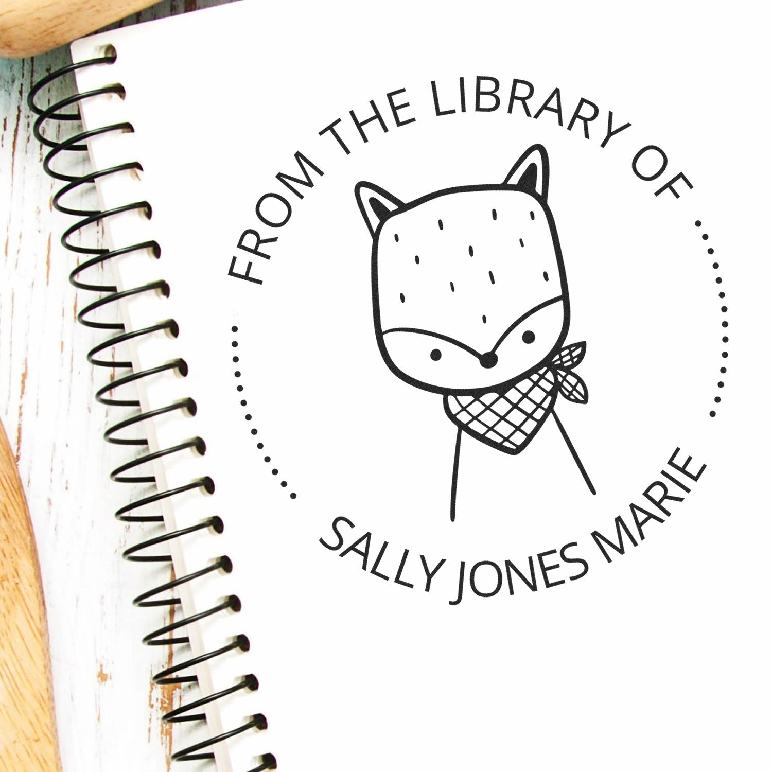 Slim Pre-Inked SnickerSnack Cartoon Animal Custom From The Private Library Of Stamp on a spiral notebook. The Personalized Home Stamper features a cute fox with a bandana, surrounded by text, on a wooden surface.