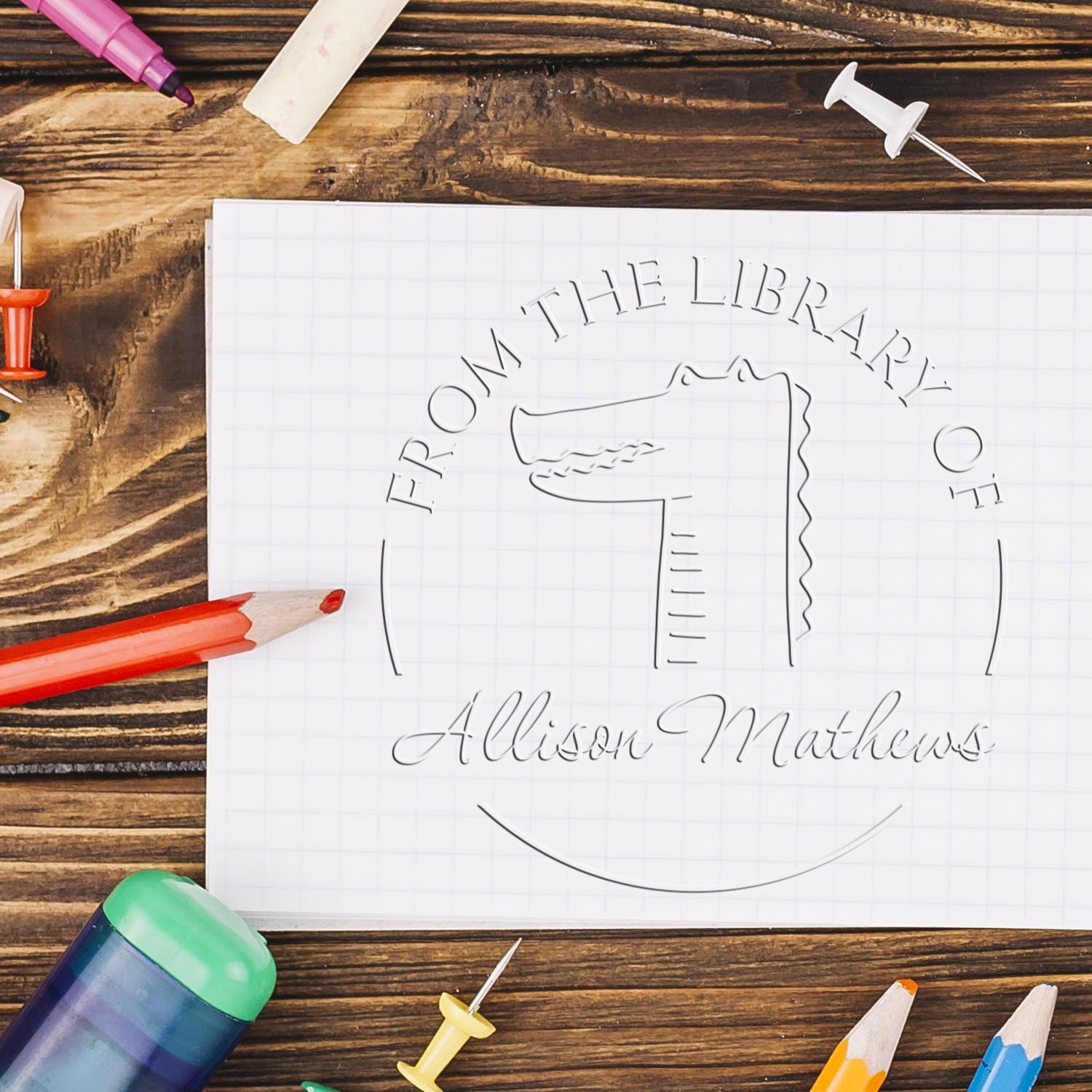 A NoodleNose cartoon animal design is embossed on paper using a personalizable book embosser. The text reads From the Library of Allison Mathews. Nearby are colorful pencils and pushpins. Custom new address stamp tools are visible on a wooden desk.