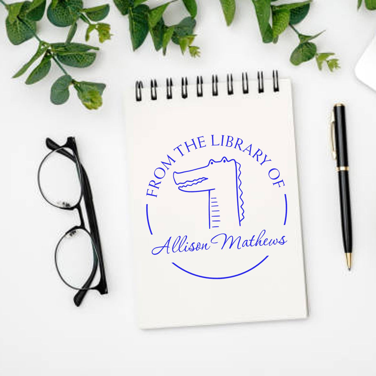 A notebook with a Slim Pre-Inked NoodleNose Cartoon Animal Custom Book Plate Stamp design featuring a crocodile and the text "From the Library of Allison Mathews." Nearby are glasses, a pen, and greenery. Ideal for a Custom-Made New Address Stamp.