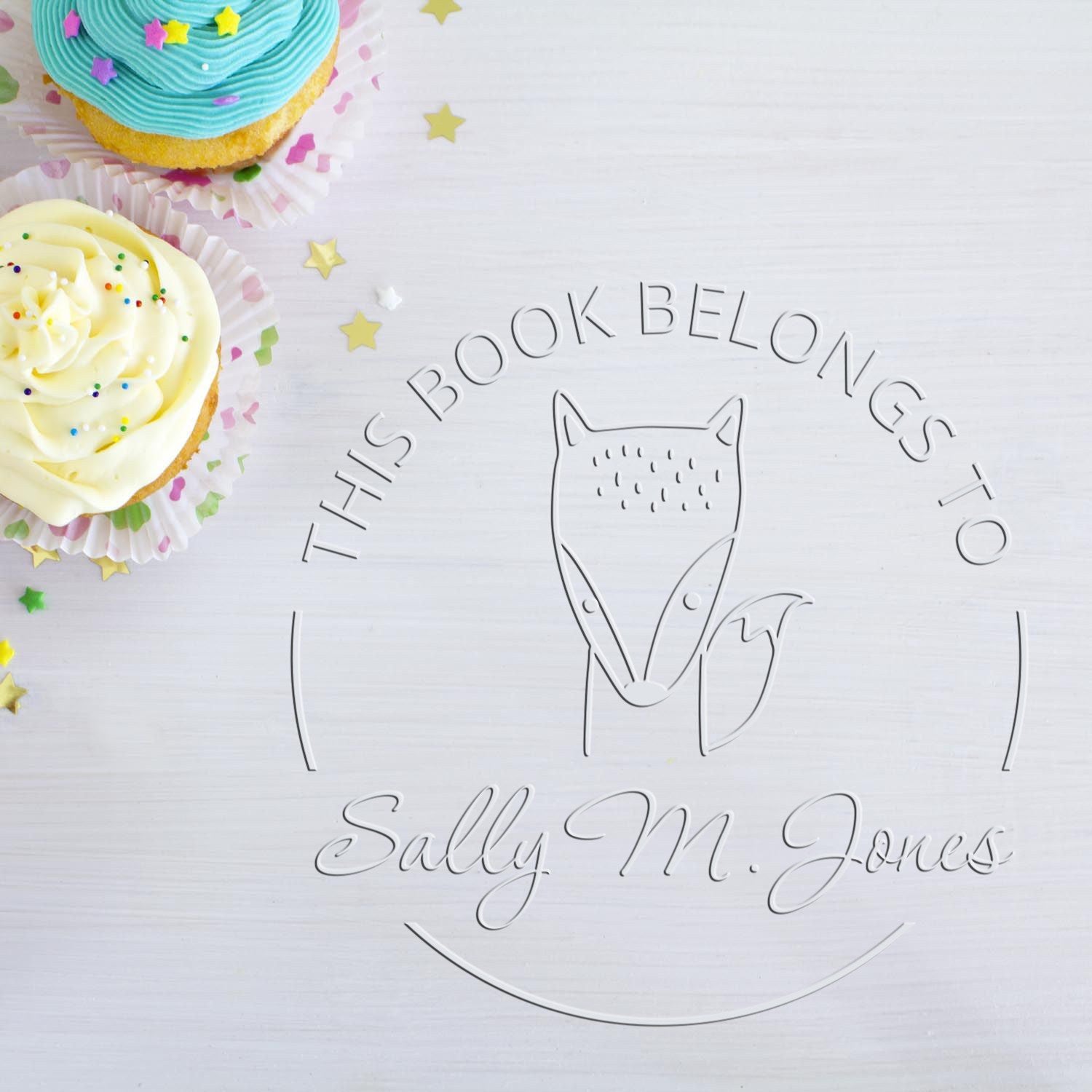 Soft BlinkBounce cartoon animal design embossed on a page with the text "This Book Belongs To Sally M. Jones." The custom-made book branding embosser is shown alongside colorful cupcakes. Includes a customized return mail stamper.