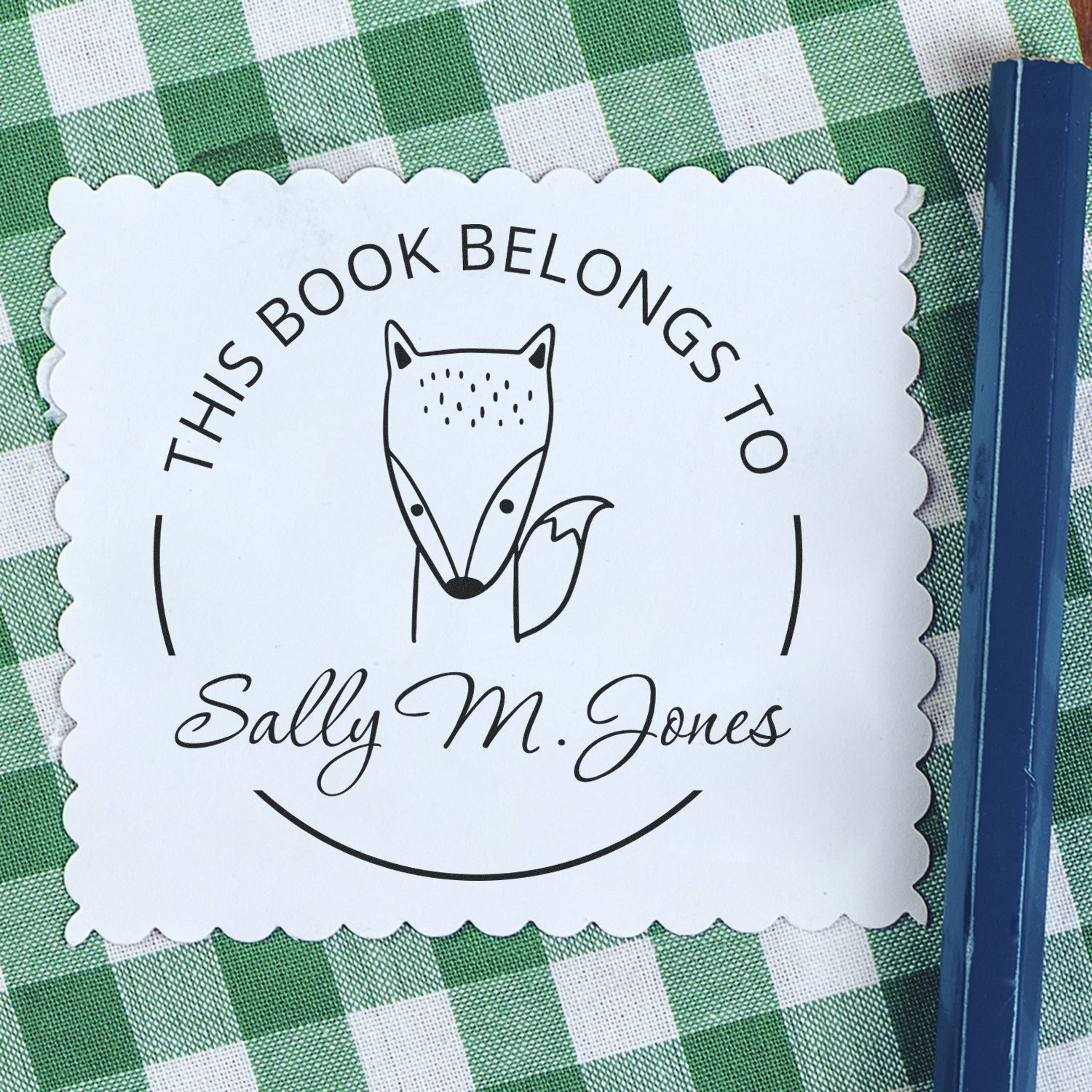 A Slim Pre-Inked BlinkBounce Cartoon Animal Custom Library Book With Name Stamp is shown on a green checkered surface. The stamp features a fox illustration and the text "This Book Belongs To Sally M. Jones." A Customizable Return Mail Stamper is beside it.