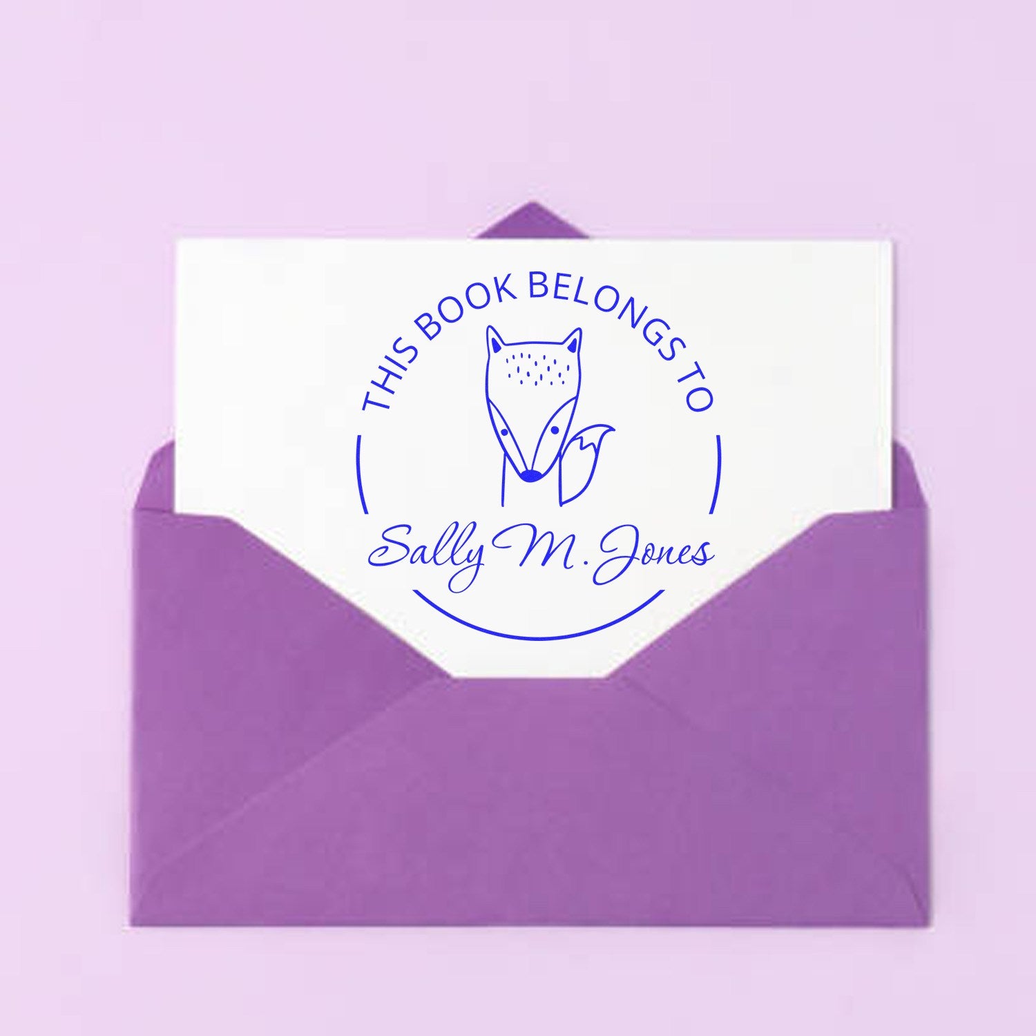 A purple envelope contains a card with a Slim Pre-Inked BlinkBounce Cartoon Animal Custom Library Book With Name Stamp design, featuring a fox and the text "This Book Belongs To Sally M. Jones." Ideal for a Customizable Return Mail Stamp.