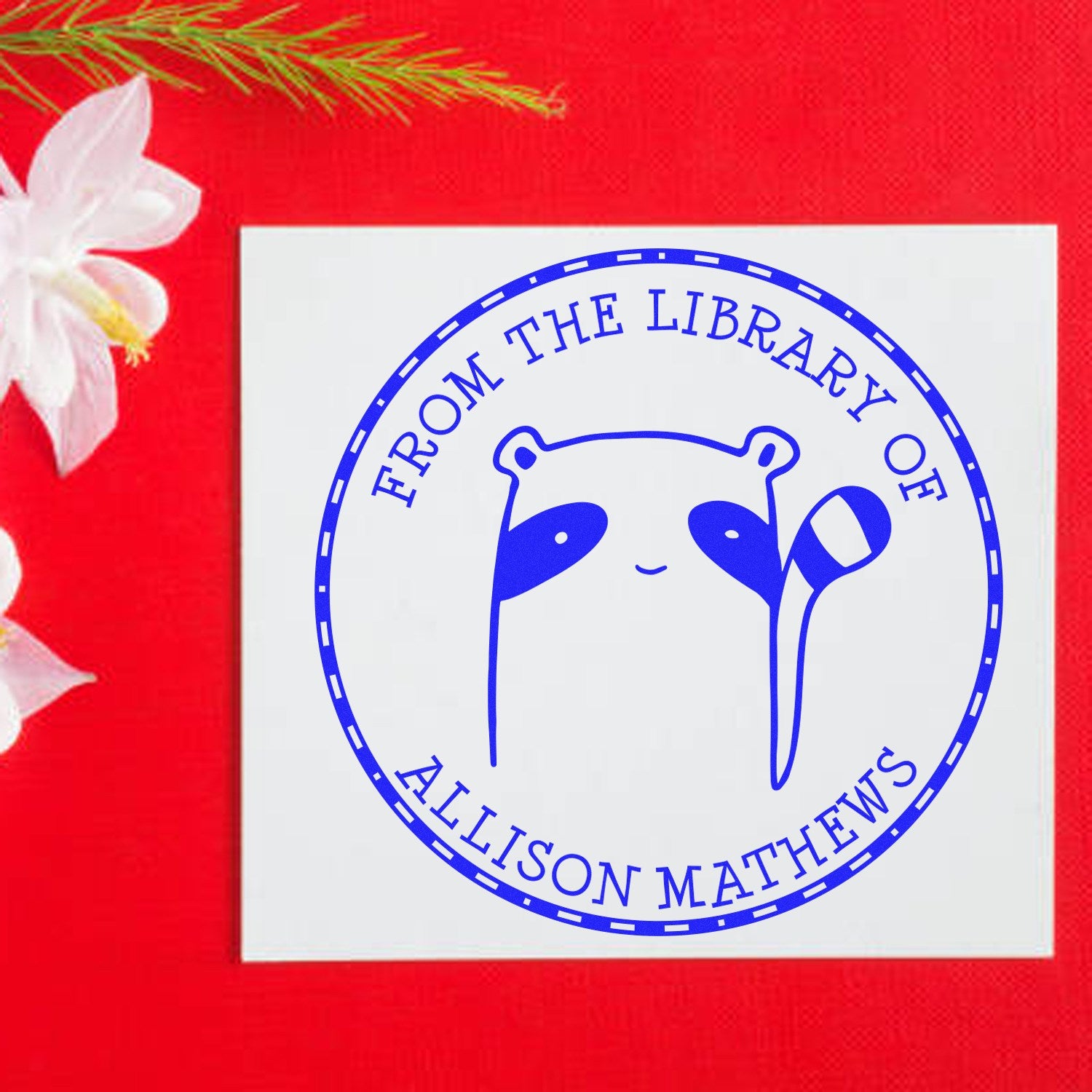 FreckleFlop cartoon animal pre-inked customizable stamp on a white card with text "From the Library of Allison Mathews" in blue. The card is placed on a red surface with white flowers. Ideal for a customized address stamp.