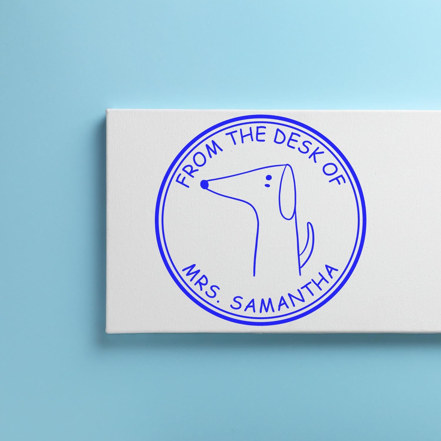 Slim Pre-Inked WiggleWag Cartoon Animal Custom From The Home Library Of Stamp on a blue background. The customizable mailing stamp features a simple cartoon dog with text "From the Desk of Mrs. Samantha" in a circular design.