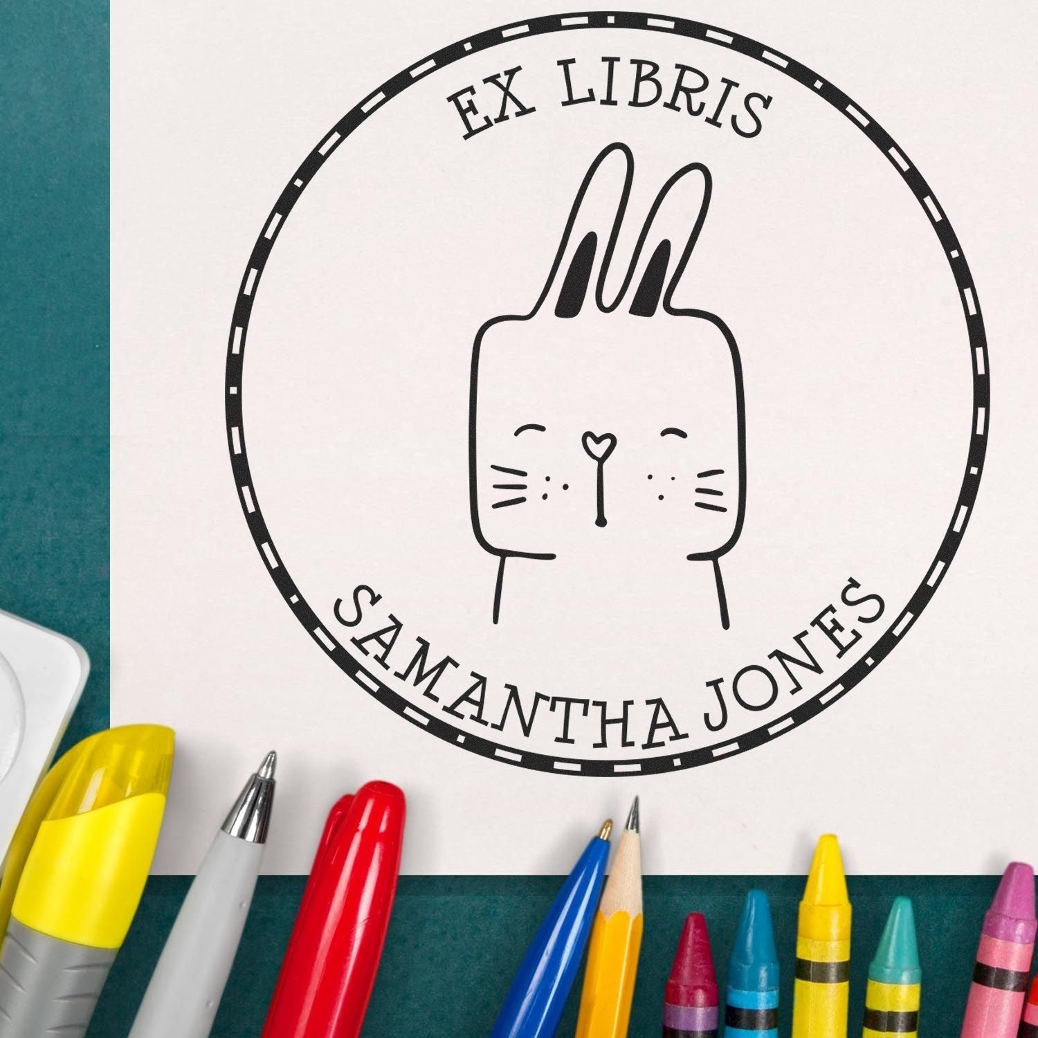 Cartoon animal stamp with "Ex Libris" and "Samantha Jones" text. Features SquirmSquiggle design, ideal for personalized address stamp or self-inking personalizable "From The Desk Of" stamp. Surrounded by colorful pens on a green surface.
