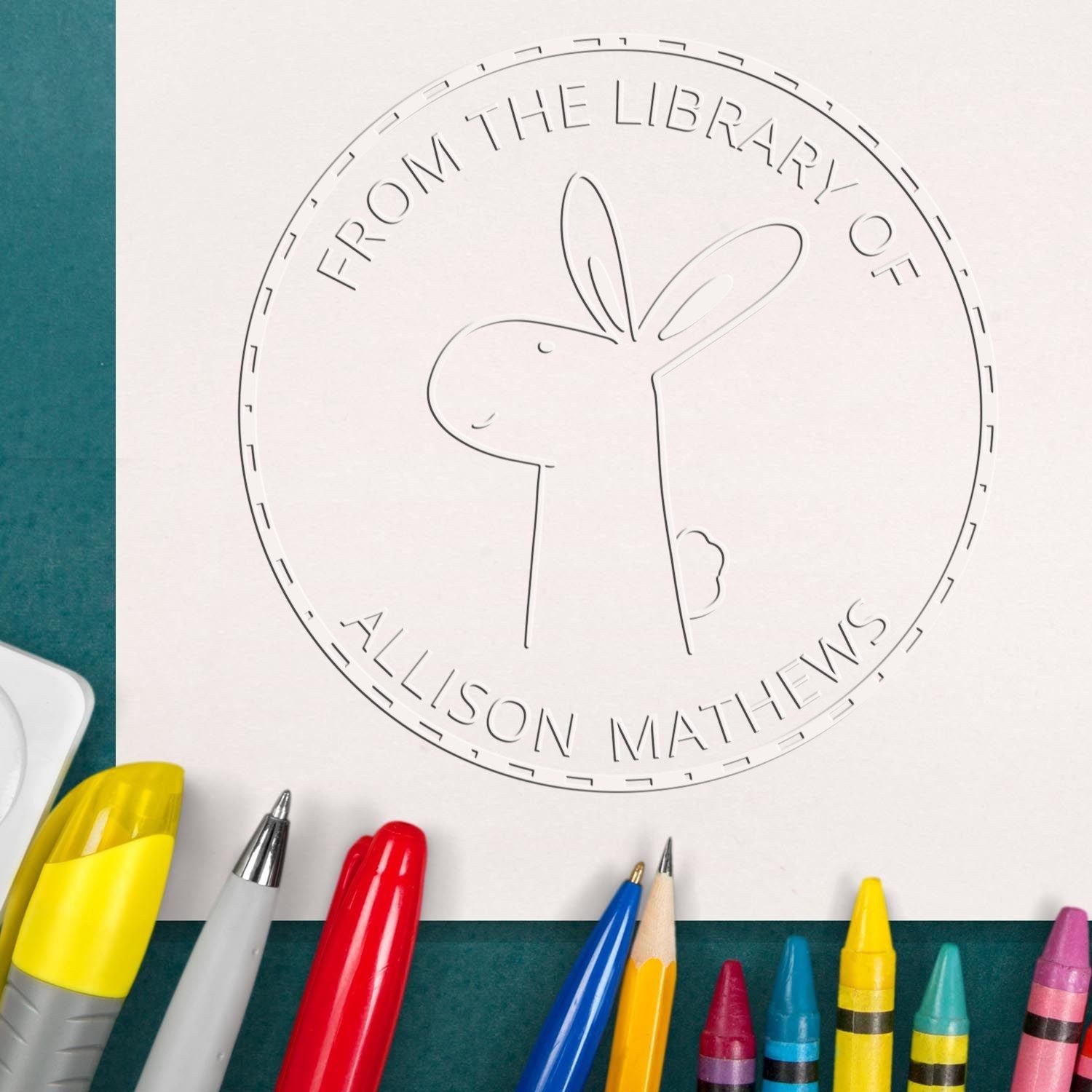 A custom-made book identification seal featuring a Soft BlinkerBelle cartoon animal, surrounded by the text "From the Library of Allison Mathews." The design is stamped on paper, with colorful pens and crayons nearby, showcasing a customized home stamp.
