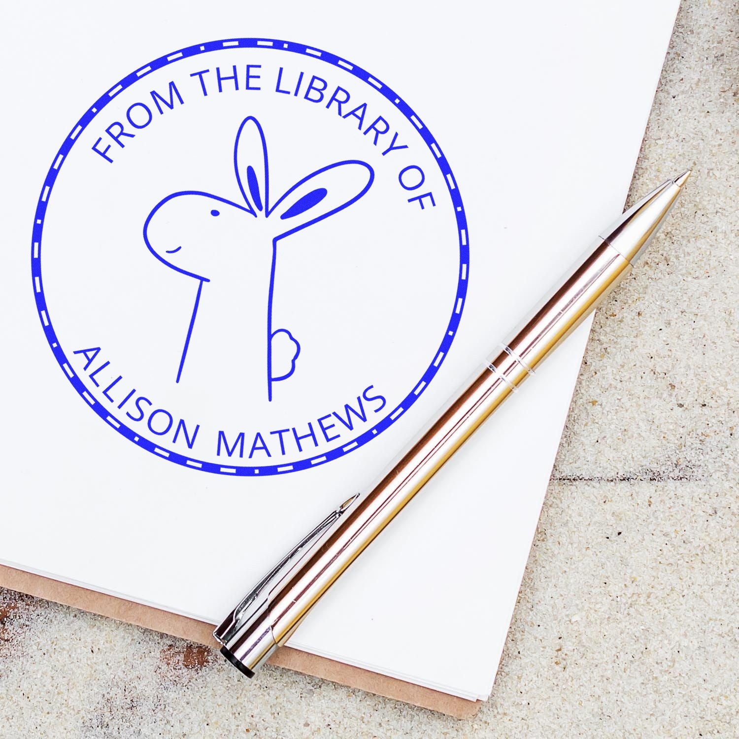A BlinkerBelle cartoon animal self-inking stamp with "This Book Belongs To" text is shown on a white page. The stamp is personalizable, featuring a custom address stamper design. A silver pen lies beside the stamped page.