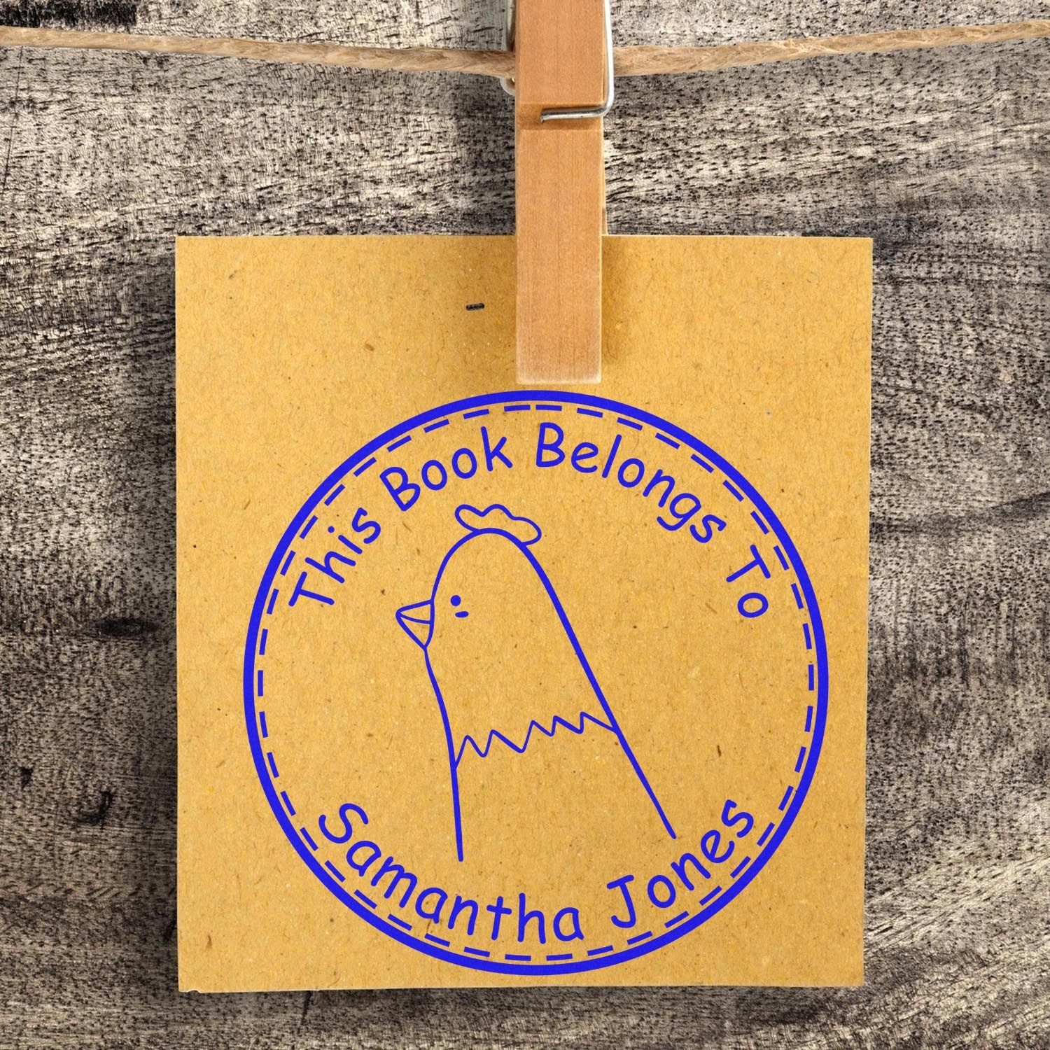 A Slim Pre-Inked FuzzleFeather Cartoon Animal Custom From The Library Of Design Stamp is shown on a brown paper, clipped to a string. The blue stamp features a cartoon bird and the text This Book Belongs To Samantha Jones. It's a Customized Mailing Stamp.