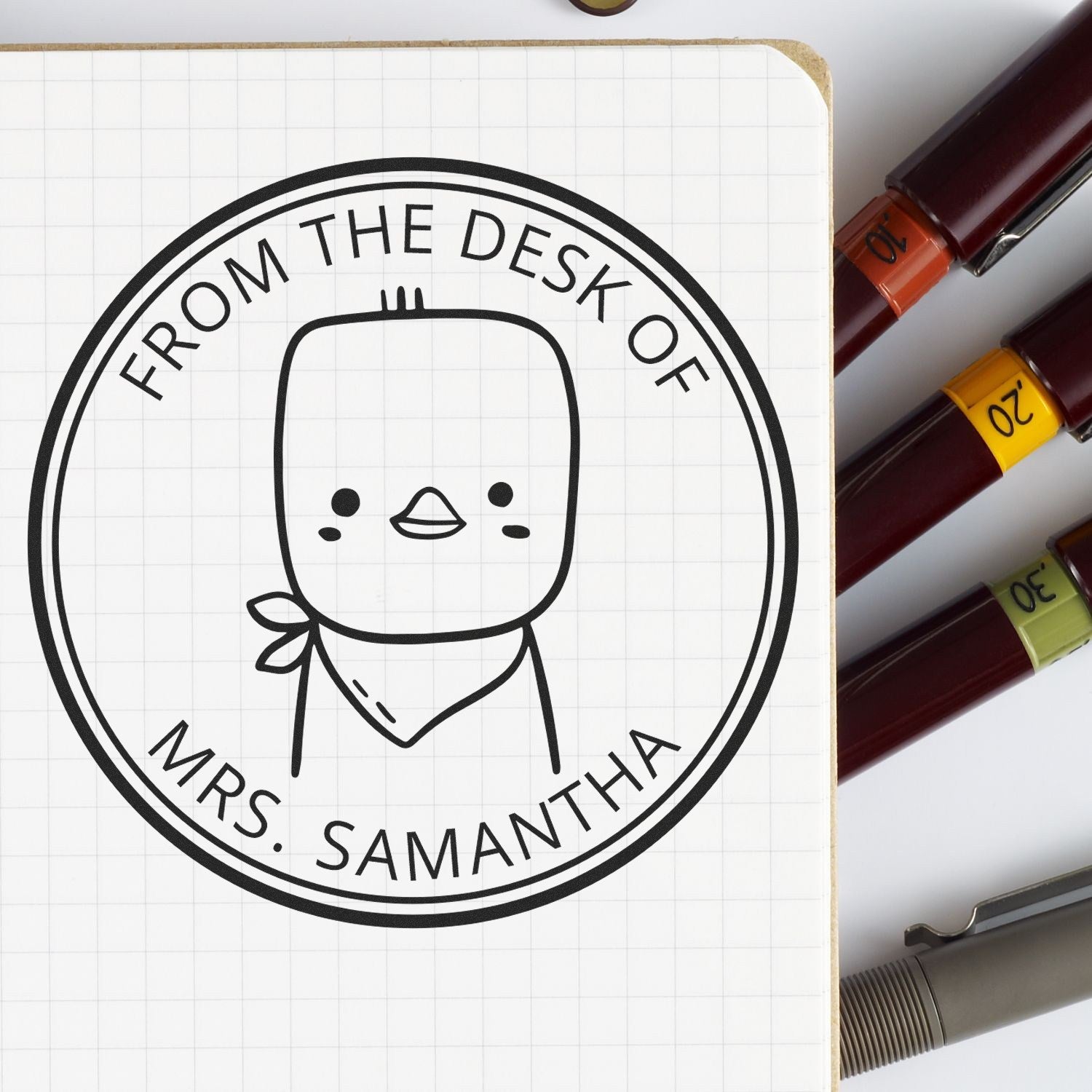 Cartoon animal stamp with "From the Desk of Mrs. Samantha" text, featuring PuddlePaddle design. Self-inking, personalizable for library books or as a custom-made home stamp. Pens are placed beside the stamp on a notebook.