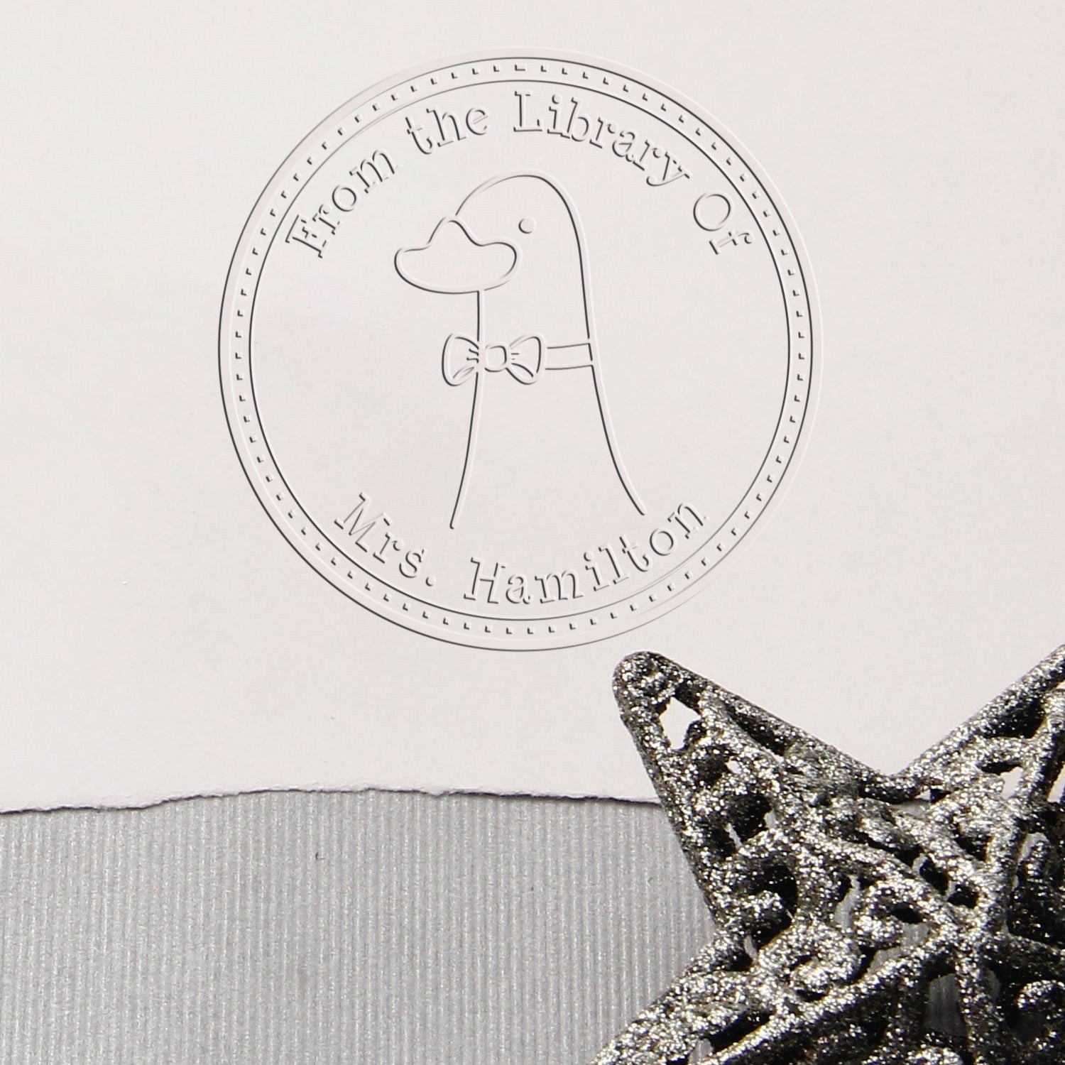 Embossed seal featuring a DoodleDuck cartoon animal with a bow tie, labeled "From the Library Of Mrs. Hamilton." The image also includes a silver star decoration. Keywords: Handheld Personalized Book Belongs To Embossing Seal, Customized Address Stamp.